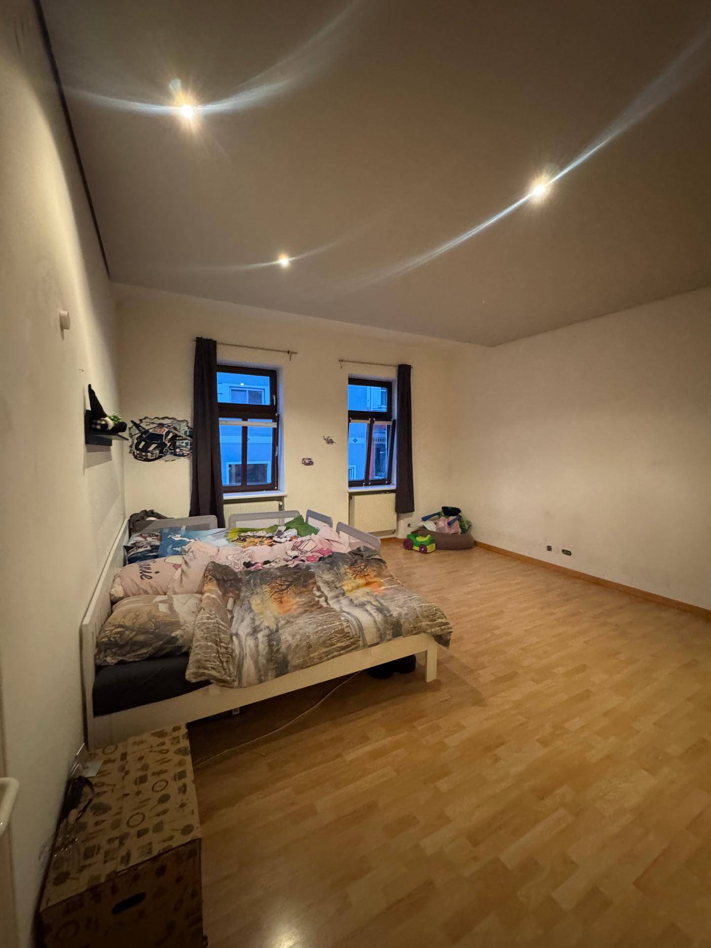 Helles Schlafzimmer mit Parkettboden, zwei Fenstern und einfacher Einrichtung.