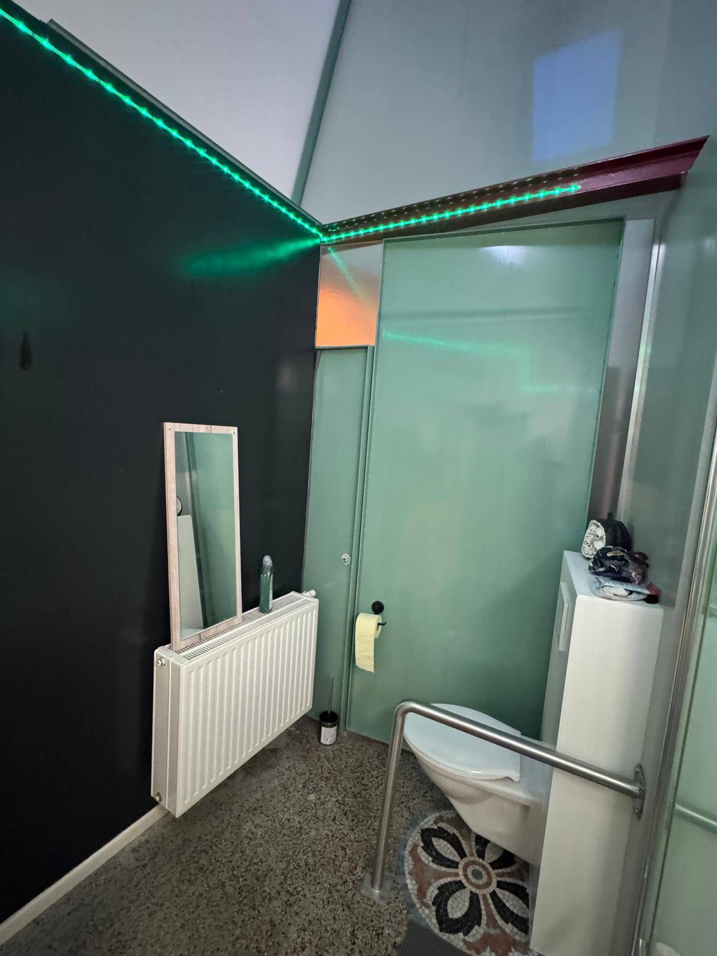Separater WC-Bereich mit Heizkörper, kleinem Spiegel und grünen LED-Lichtern an der Wand.