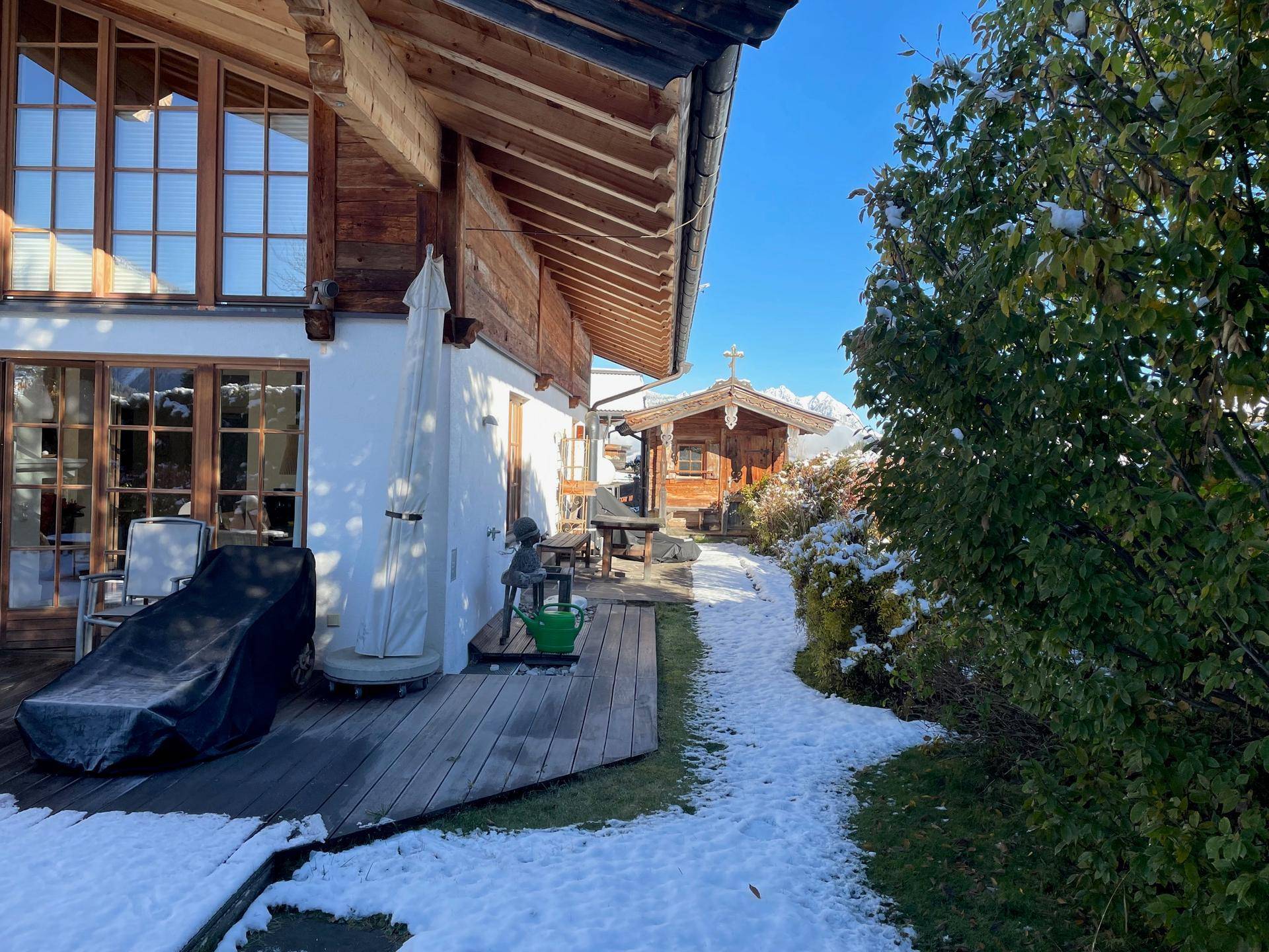 Holzterrasse am Haus mit Liegestuhl und einem verschneiten Pfad, der zu einem Holzhäuschen führt.
