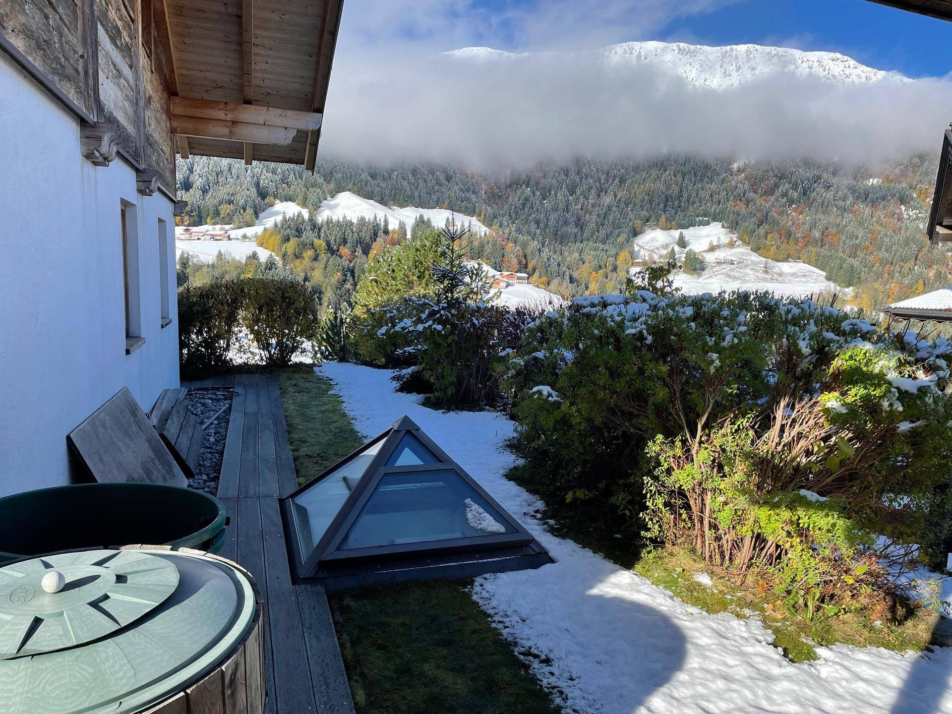 Gartenbereich mit Holzterrasse, Schneeresten und Blick auf die verschneiten Berge im Hintergrund.