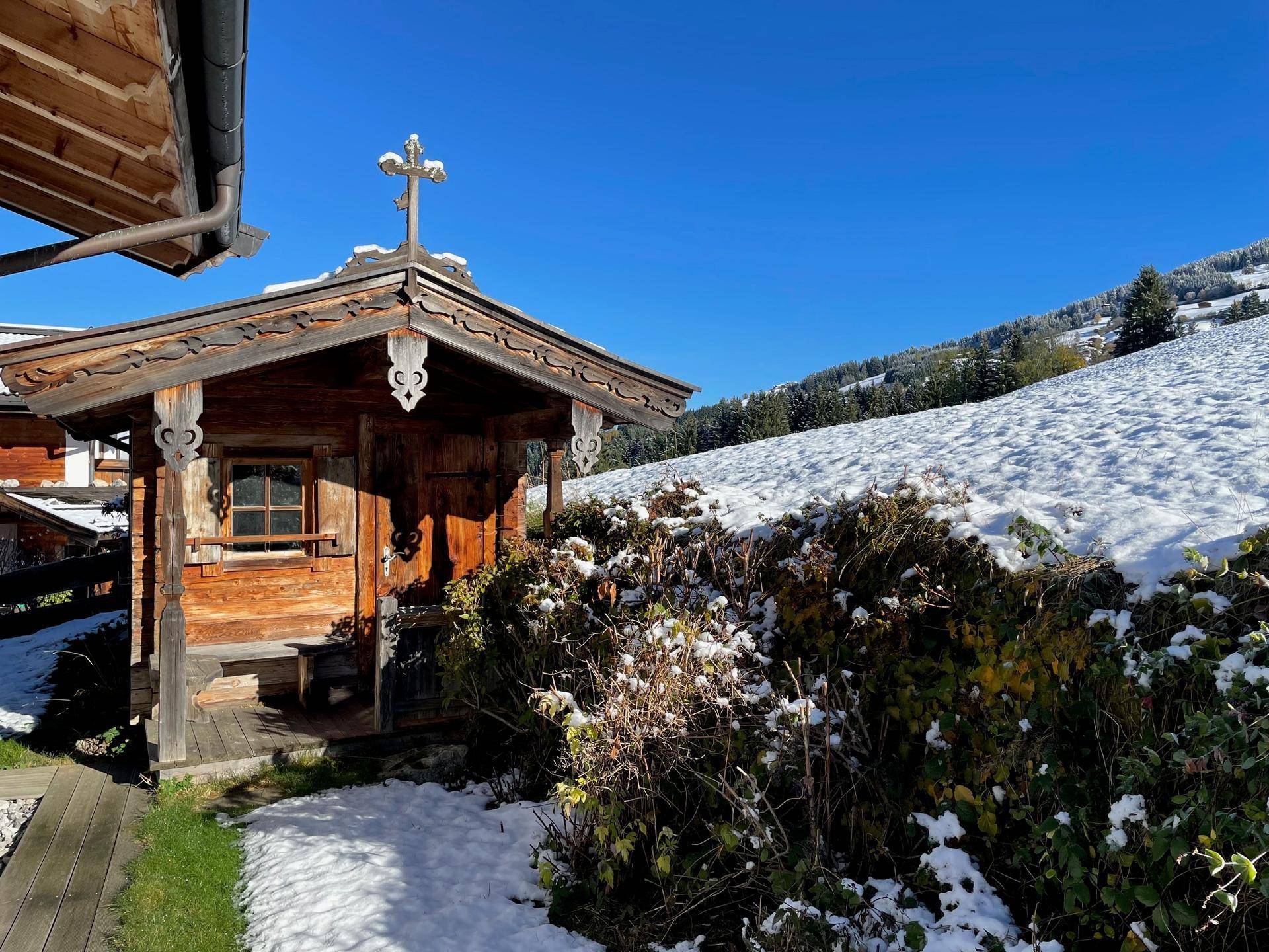 Traditionelles Holzhäuschen mit Kreuz im verschneiten Garten und sonnigem Himmel.