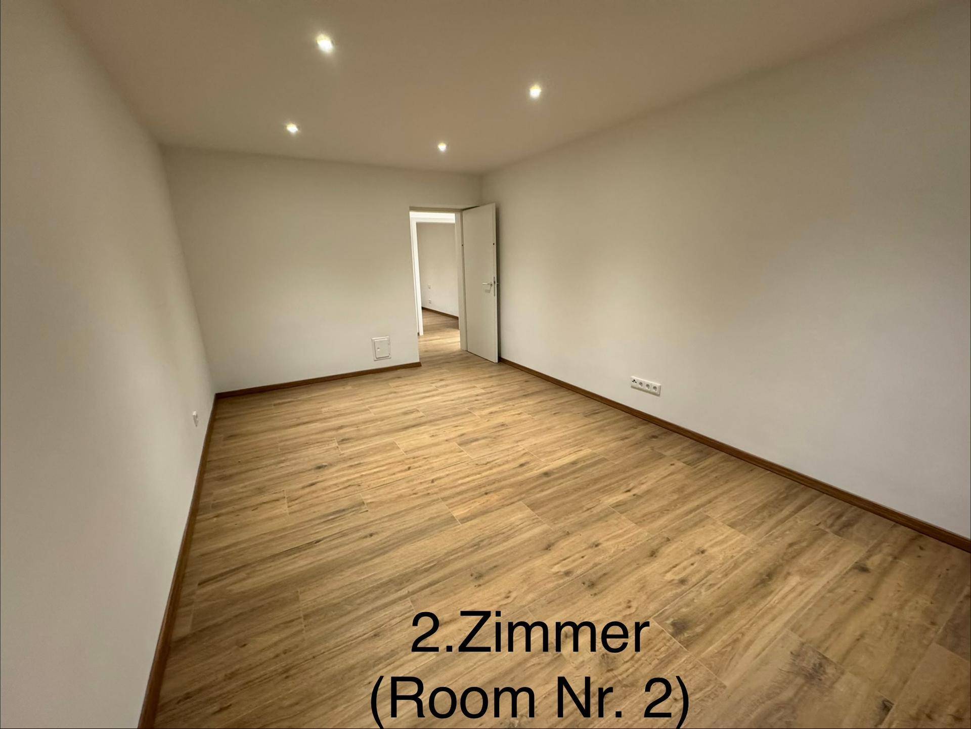 Geräumiges Zimmer mit Holzboden, weißen Wänden und Zugang zu einem weiteren Raum.