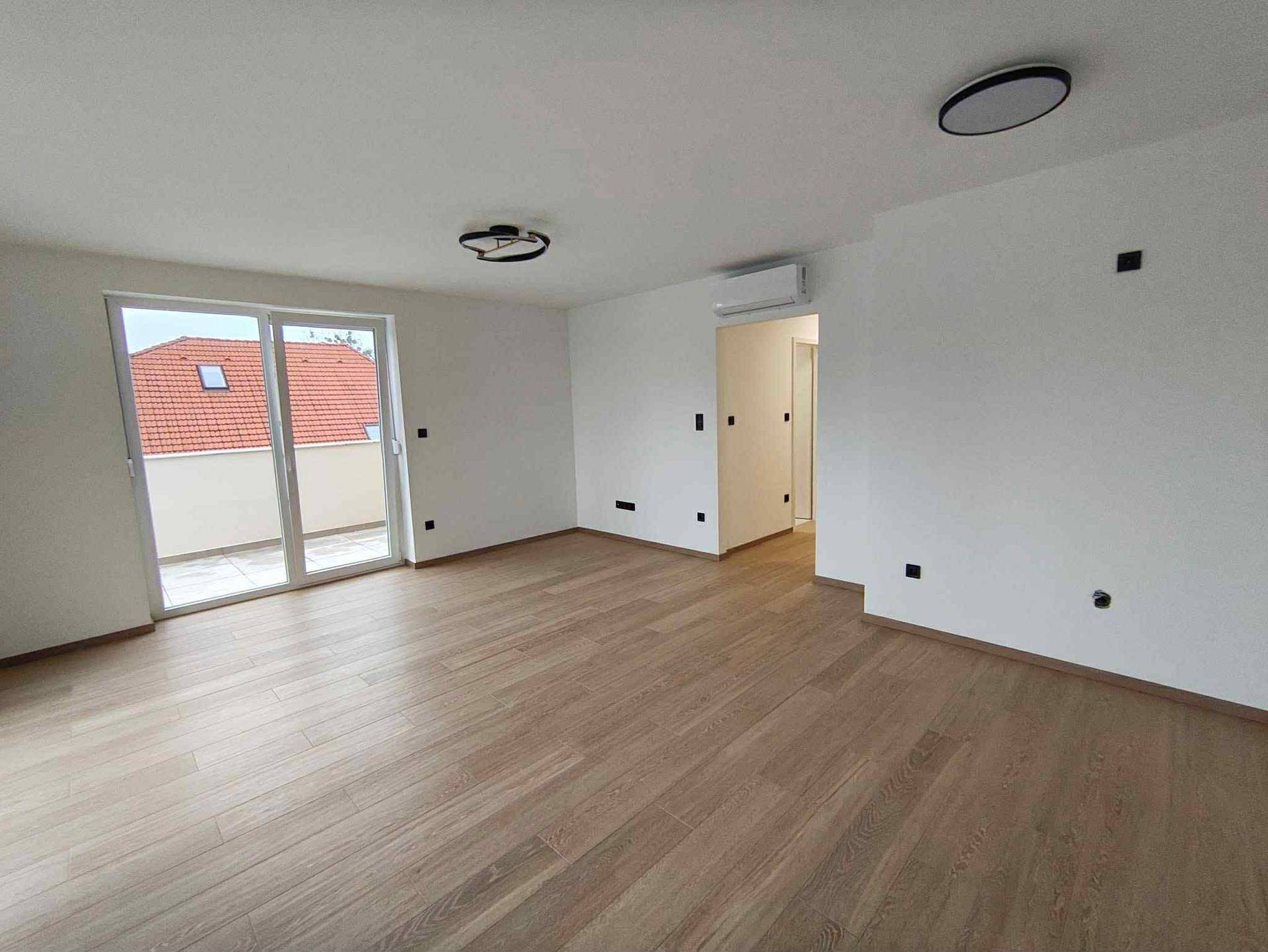 Helles, leeres Wohnzimmer mit Holzboden und großem Schiebefenster zum Balkon.