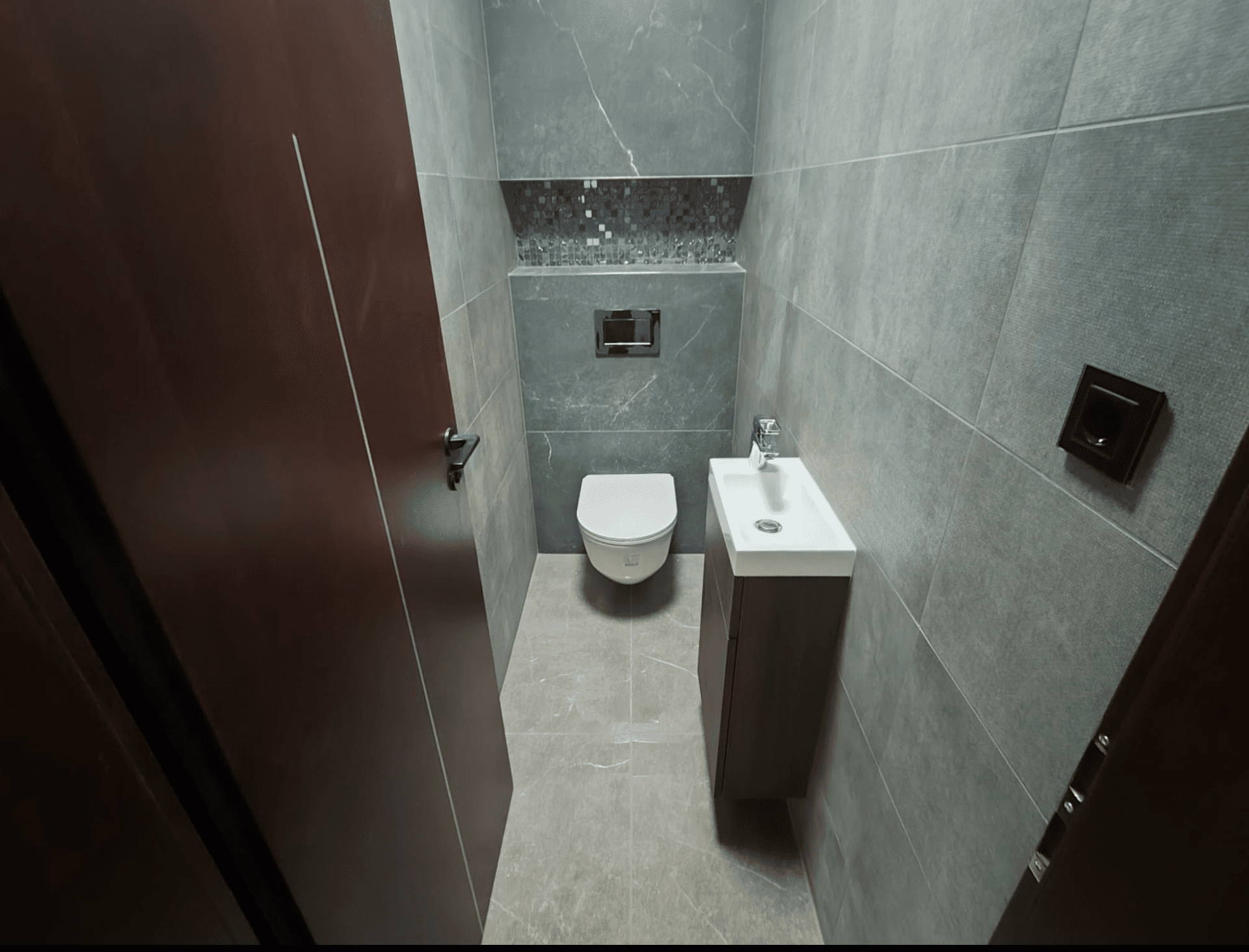 Kompaktes Gäste-WC mit wandhängendem WC, kleinem Waschbecken und dunklen Fliesen.
