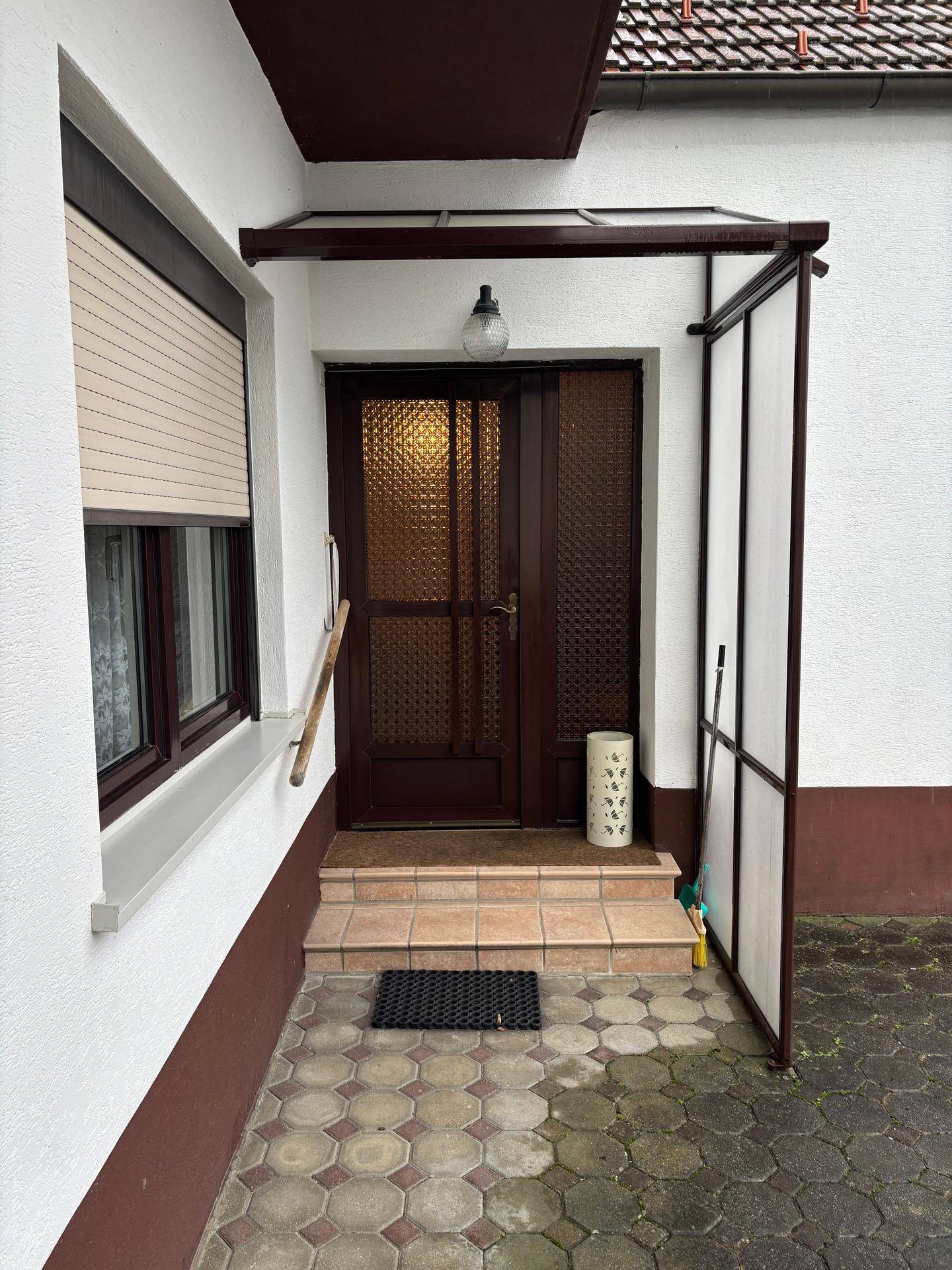 Eingangsbereich mit brauner Holztür, Vordach und kleinen Stufen, umgeben von gepflastertem Boden.