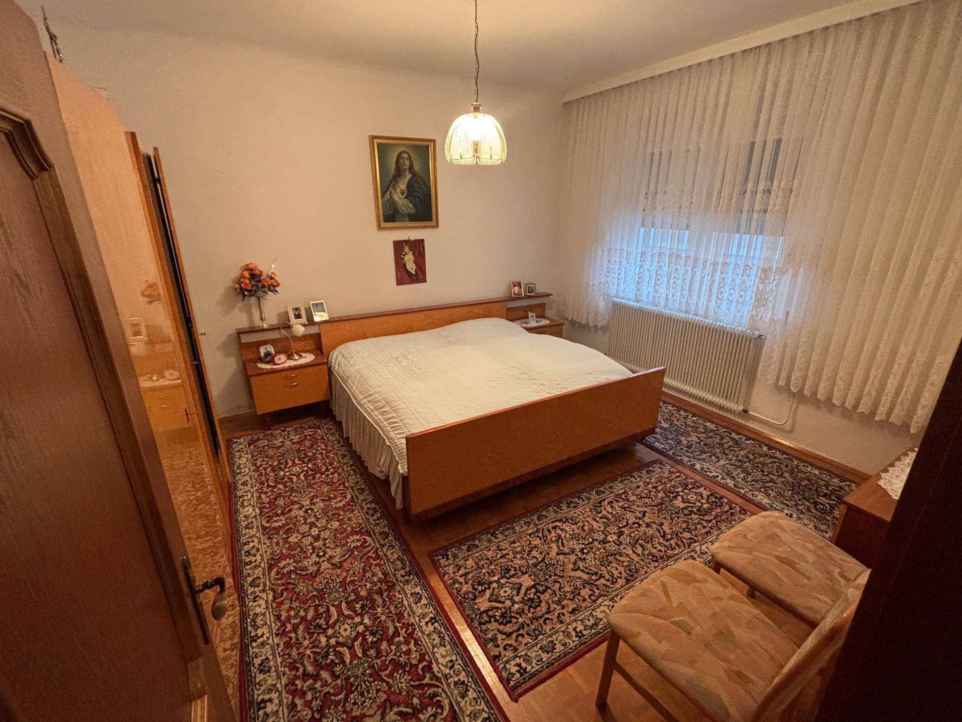 Schlafzimmer mit Doppelbett, Teppichläufern und einem Fenster mit Vorhängen in hellem Ambiente.