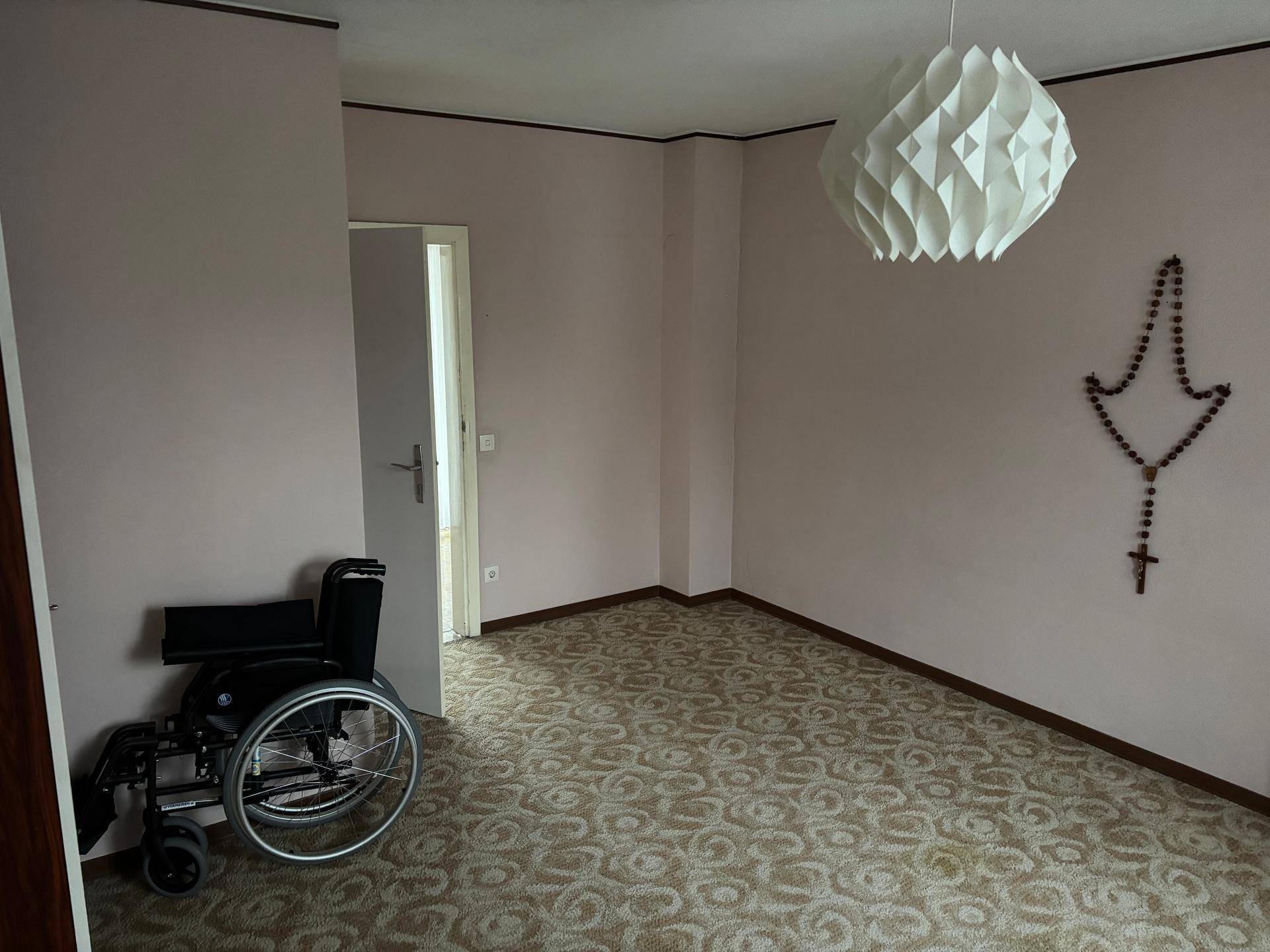 Geräumiges Zimmer mit Teppichboden, rosa Wänden und einem Rollstuhl, der Barrierefreiheit andeutet.