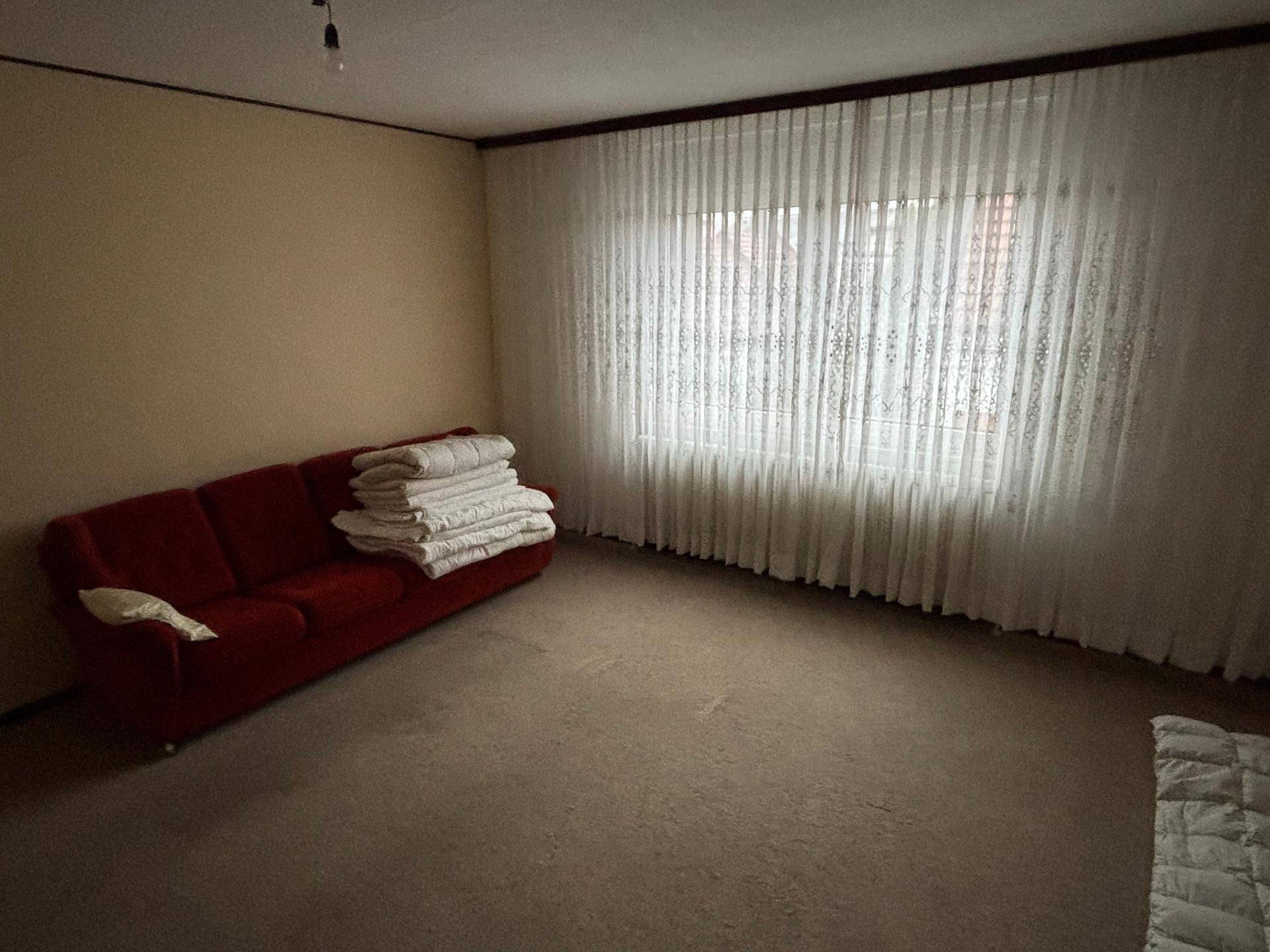 Helles Zimmer mit Teppichboden, rotem Sofa und einem großen Fenster mit weißen Vorhängen.