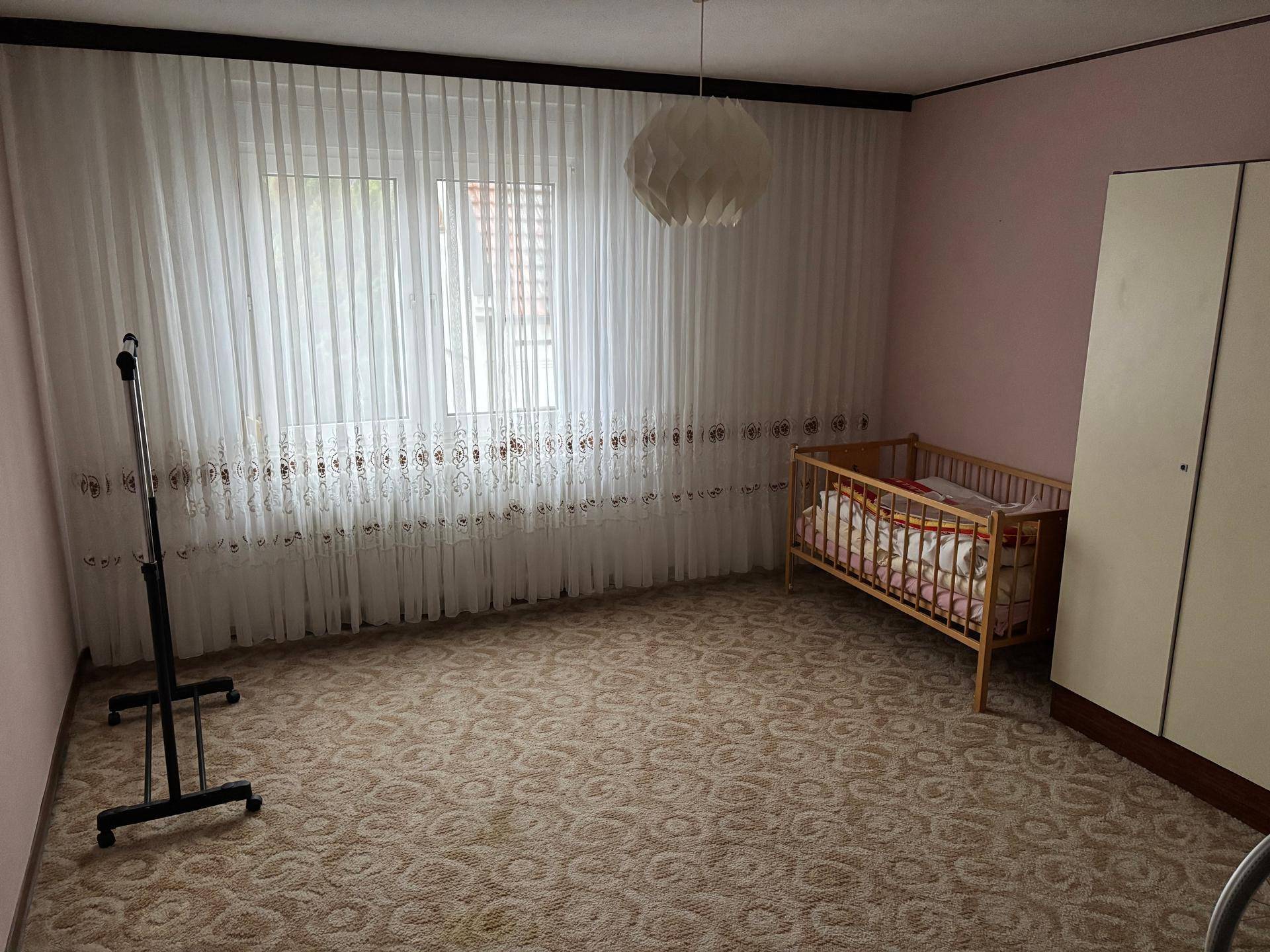 Kinderzimmer oder Schlafzimmer mit Teppichboden, Babybett und einem großen Fenster mit Vorhängen.