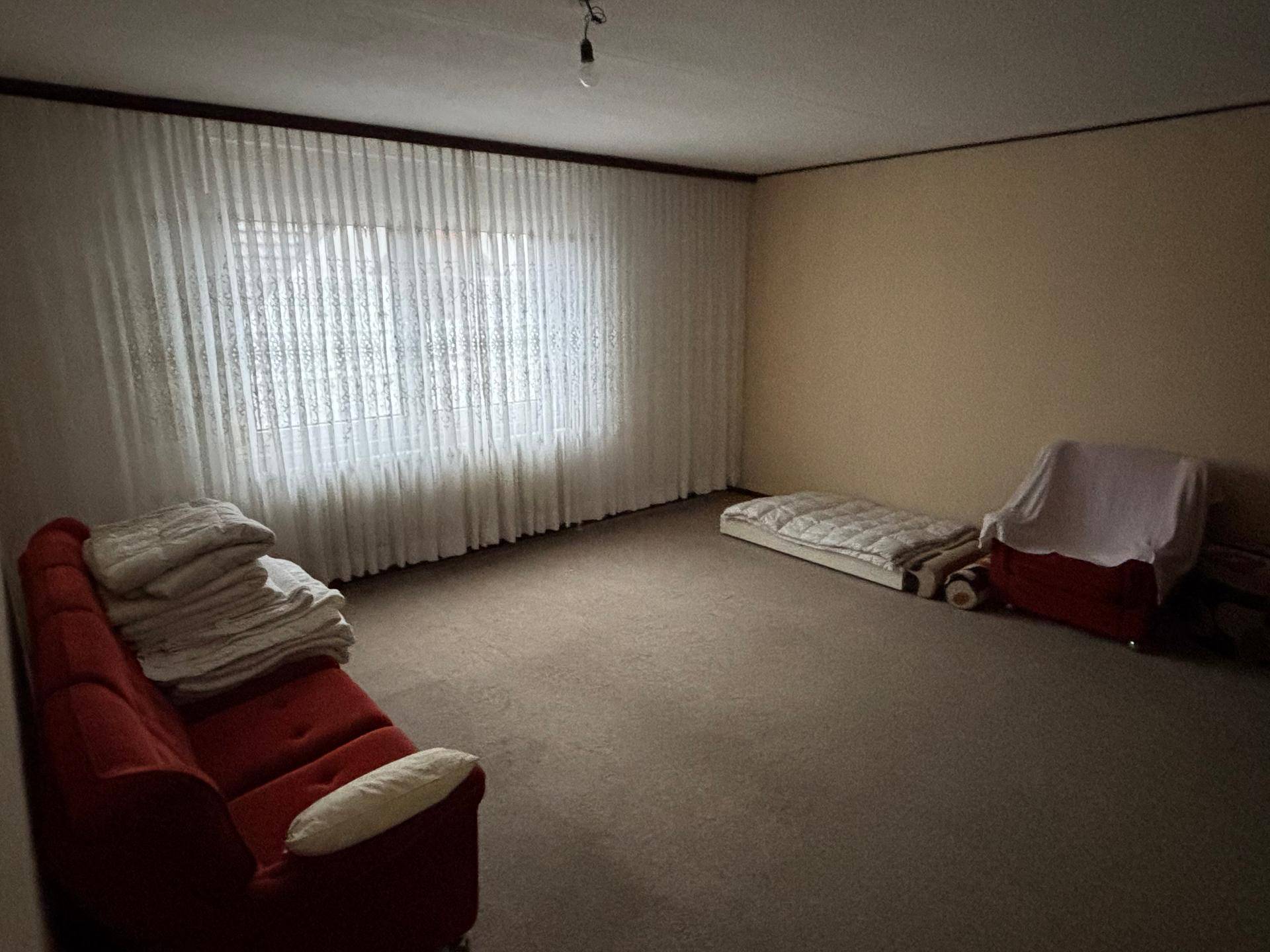 Großes Zimmer mit Teppichboden, rotem Sofa und einem Fenster, das viel Tageslicht spendet.