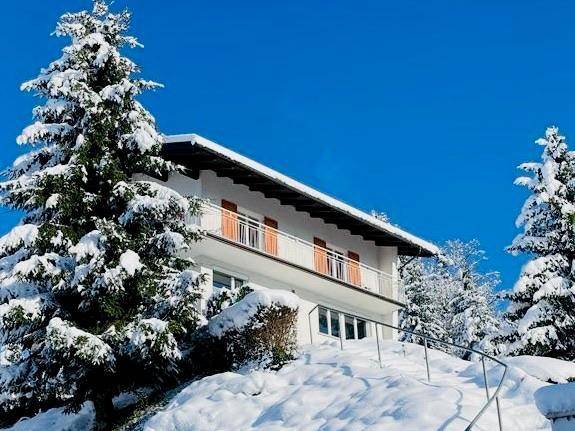 Hausfassade im Winter mit schneebedeckten Bäumen und blauem Himmel.