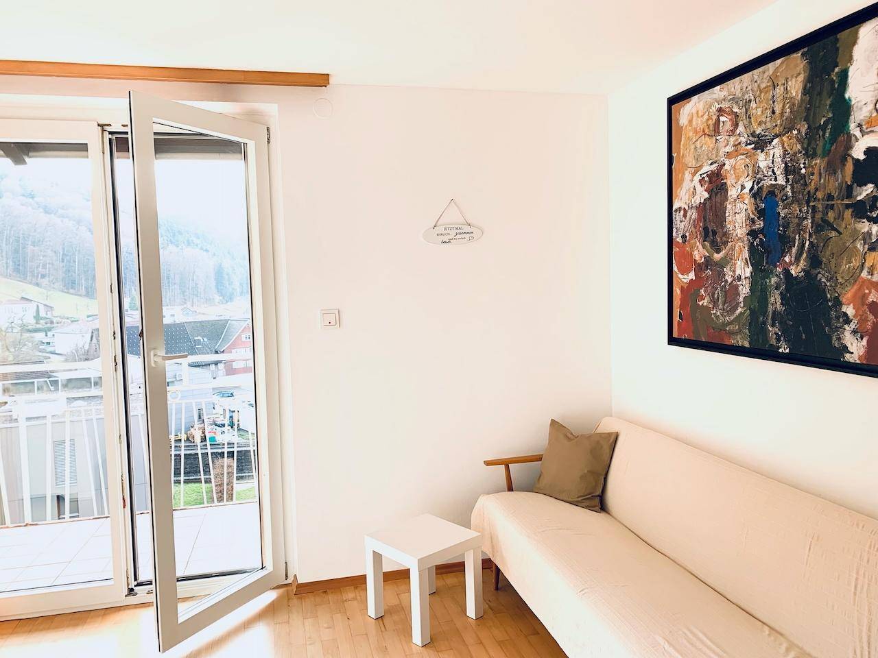 Heller Wohnbereich mit Sofa, Kunstwerk und Balkonzugang mit Blick ins Grüne.