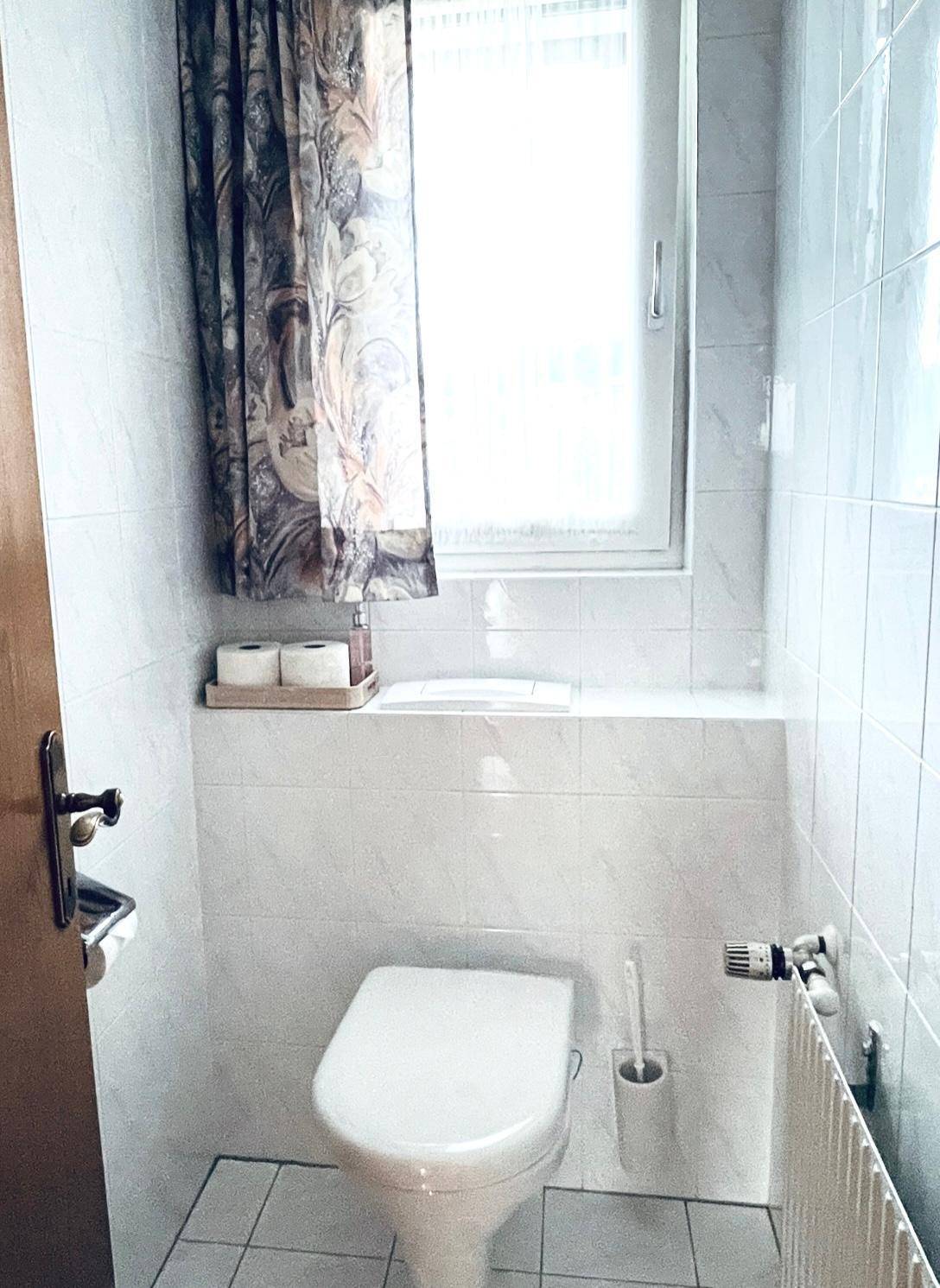 Kleines WC mit weiß gefliesten Wänden, Toilette und Fenster.