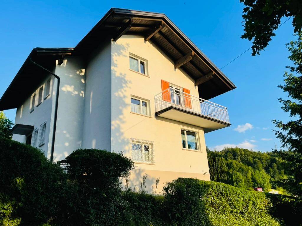 Hausfassade mit Balkon, Fensterläden und umgebender grüner Landschaft bei Sonnenuntergang.