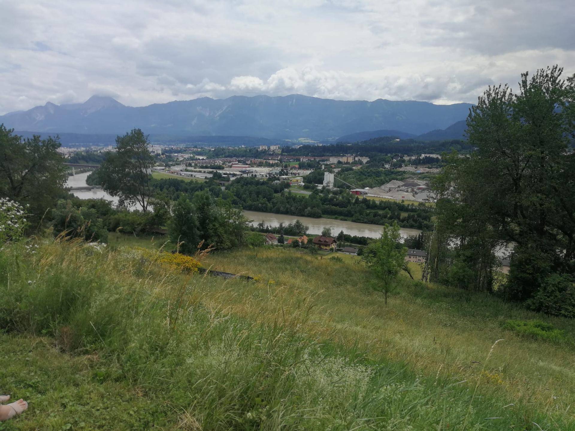 Grüne Hügellandschaft mit weitem Blick auf einen Fluss, eine Stadt und die dahinterliegenden Berge unter bewölktem Himmel.