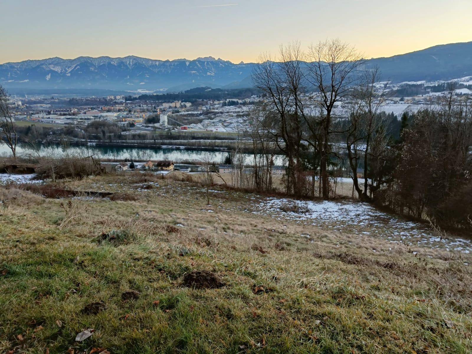Blick über eine leicht verschneite Wiese auf einen Fluss, eine Stadt und die umliegende Berglandschaft.
