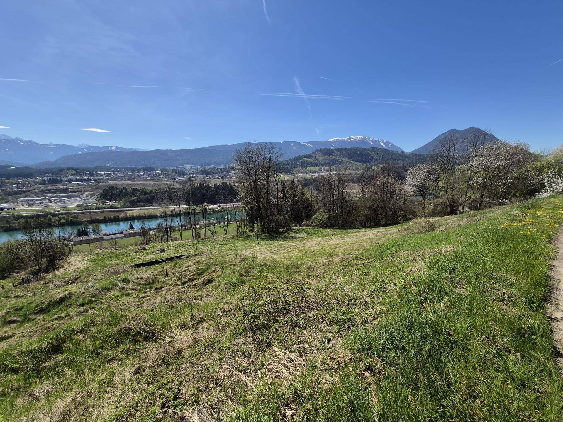 Sonniger Ausblick über eine grüne Wiese auf einen Fluss, eine Stadt und die schneebedeckten Berge im Frühling.