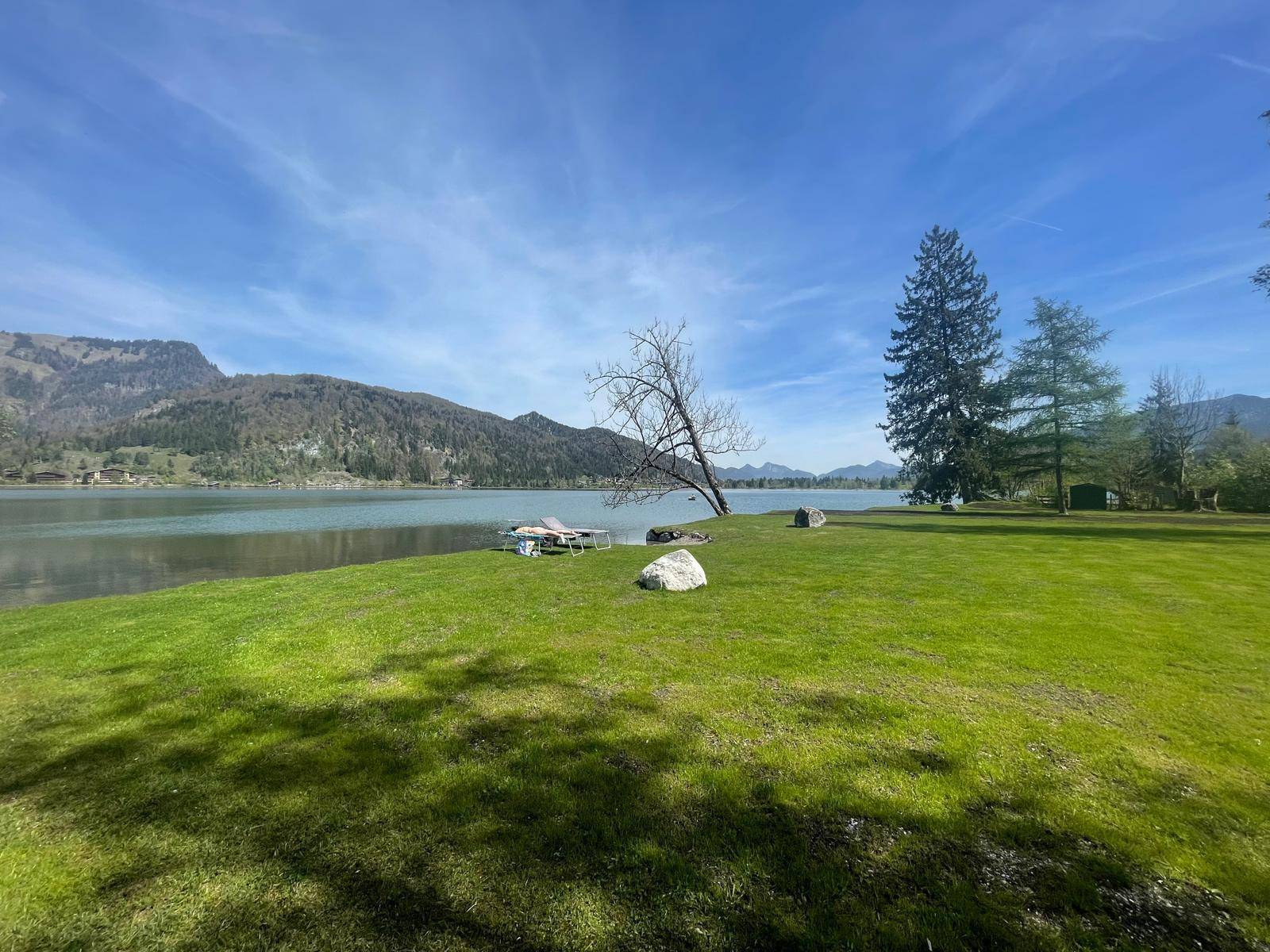 Malerische Landschaft mit einem See, grünen Wiesen und Bergen im Hintergrund unter blauem Himmel.