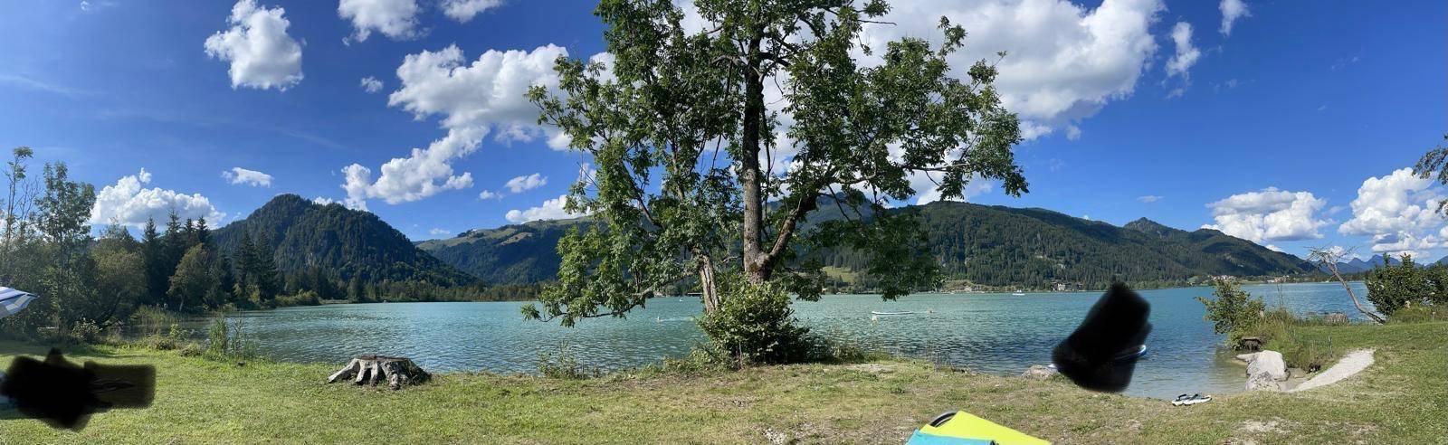 Panoramablick auf einen türkisfarbenen See umgeben von bewaldeten Bergen unter blauem Himmel.