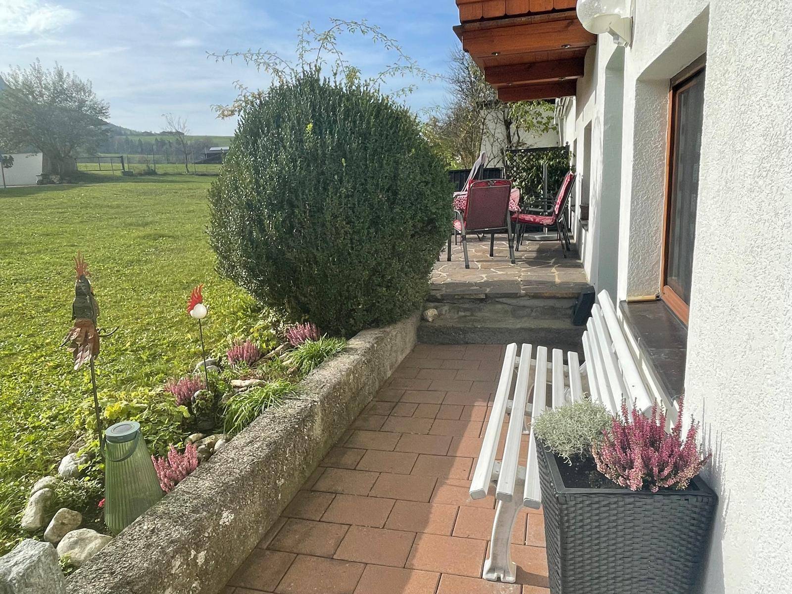 Gepflasterte Terrasse mit Sitzbank, Blumenbeeten und Blick ins Grüne.