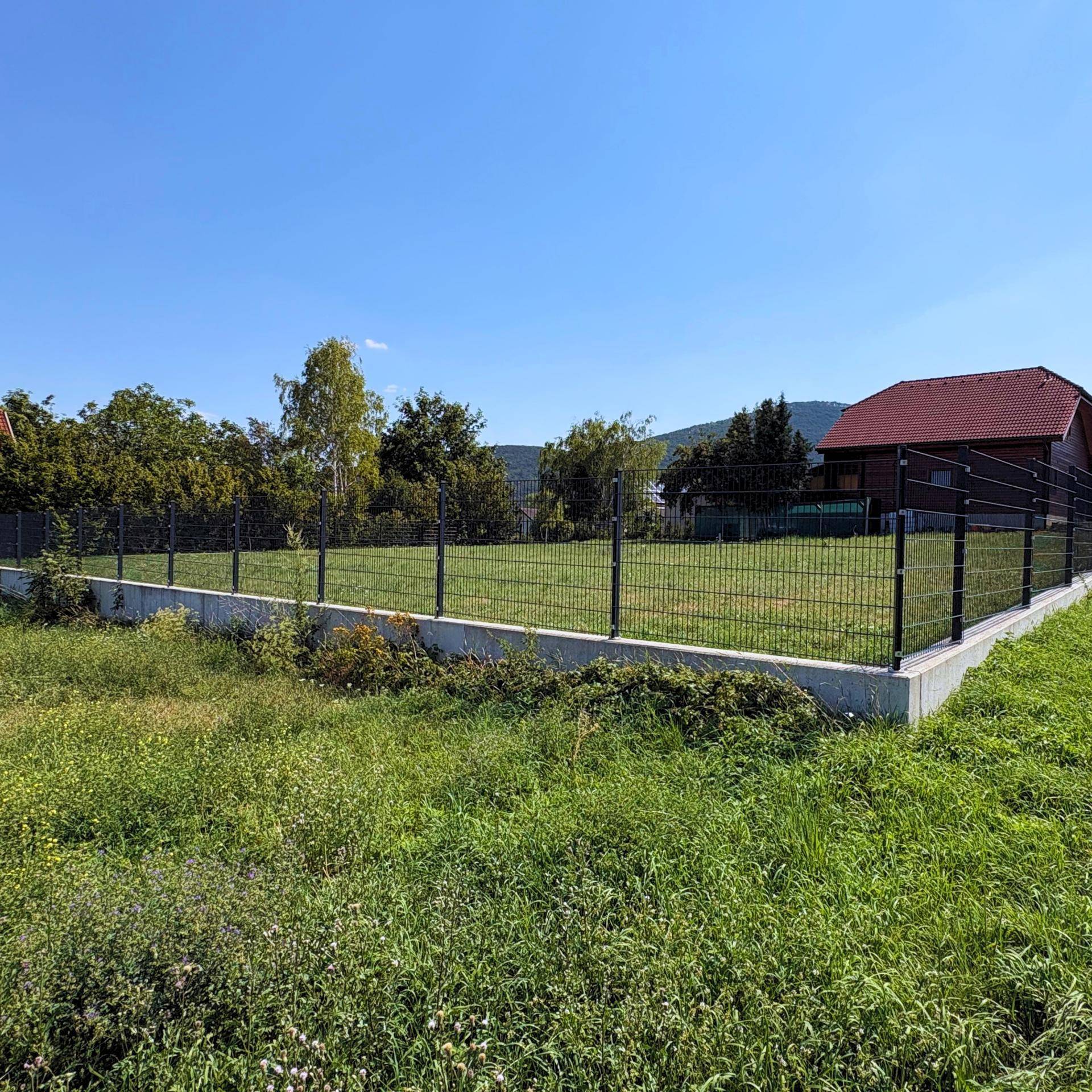 Eingezäuntes Grundstück mit grüner Wiese und Bäumen im Hintergrund unter strahlend blauem Himmel.