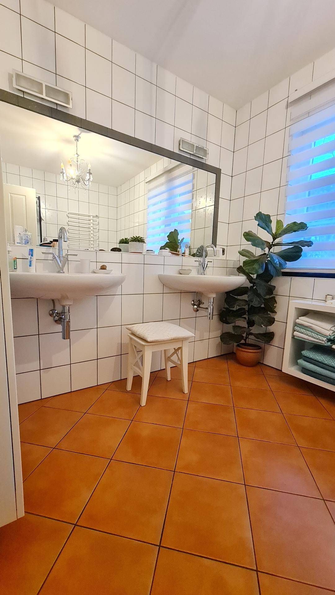 Badezimmer mit Doppelwaschbecken, großem Spiegel, weißen Fliesen und Terrakotta-Boden.
