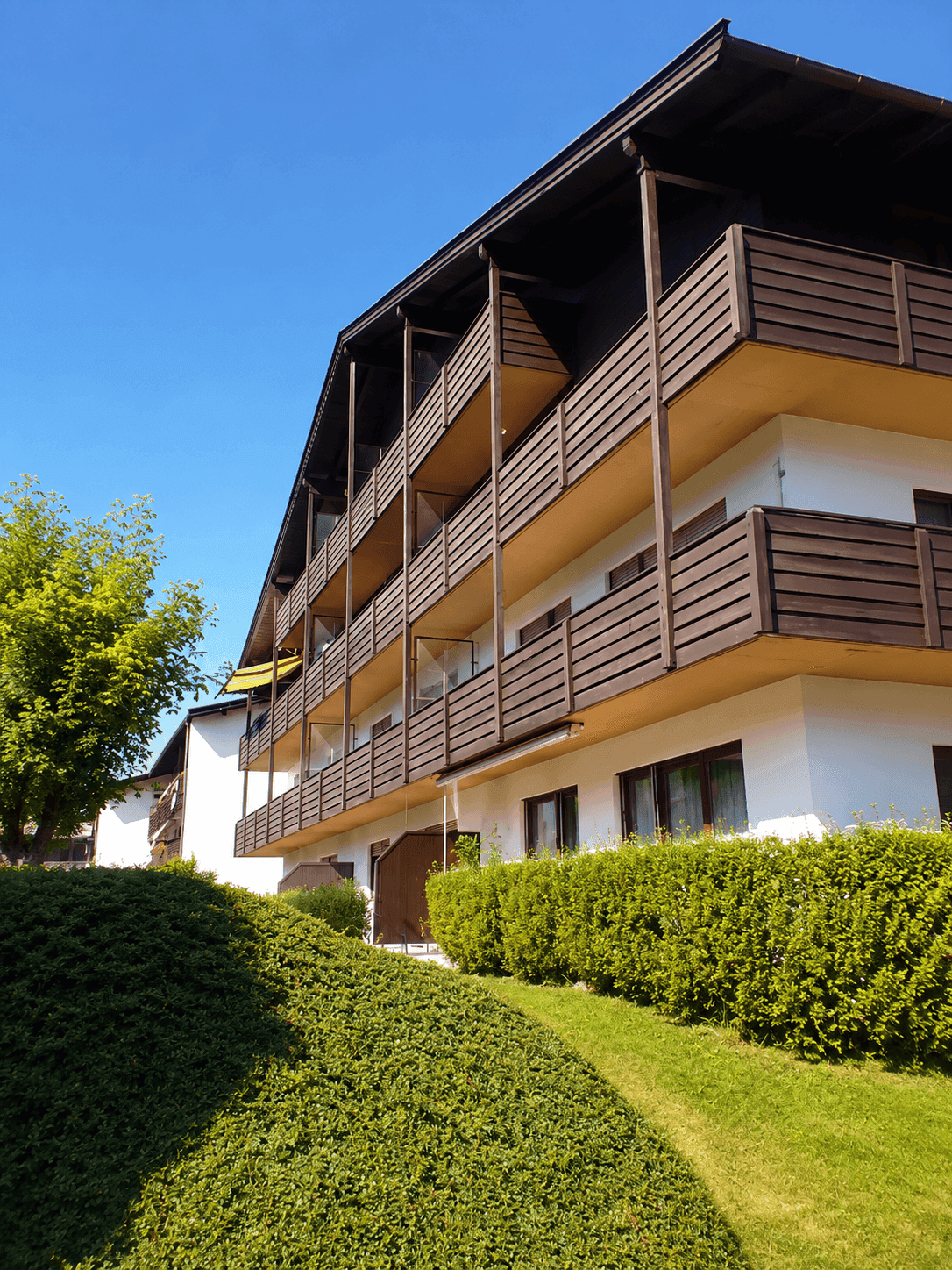 Mehrstöckiges Gebäude mit vielen Holzbalkonen und gepflegter Hecke im Vordergrund.