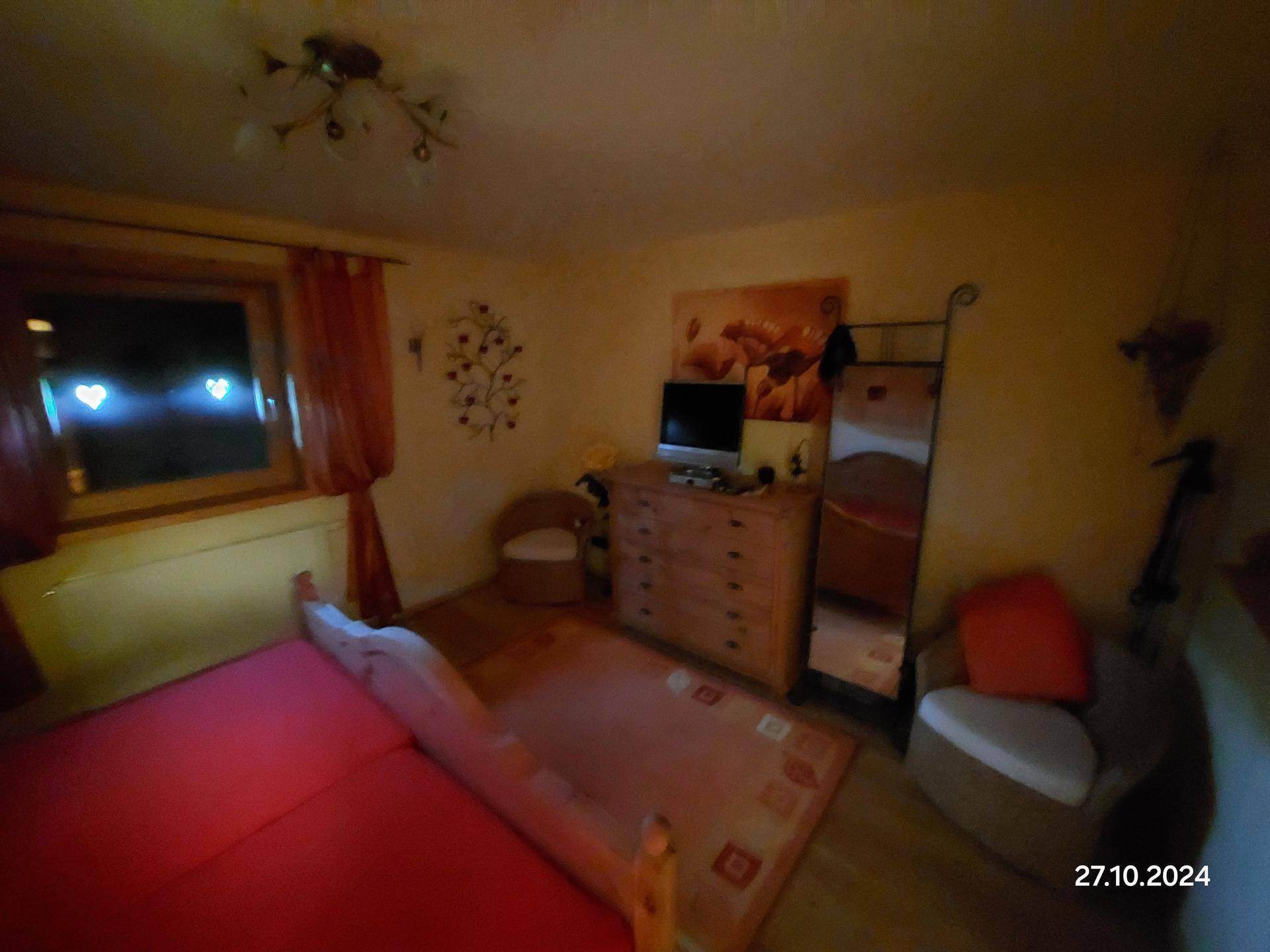 Gemütliches Schlafzimmer mit rotem Bett, Holzmöbeln und einem Fenster mit Vorhängen.
