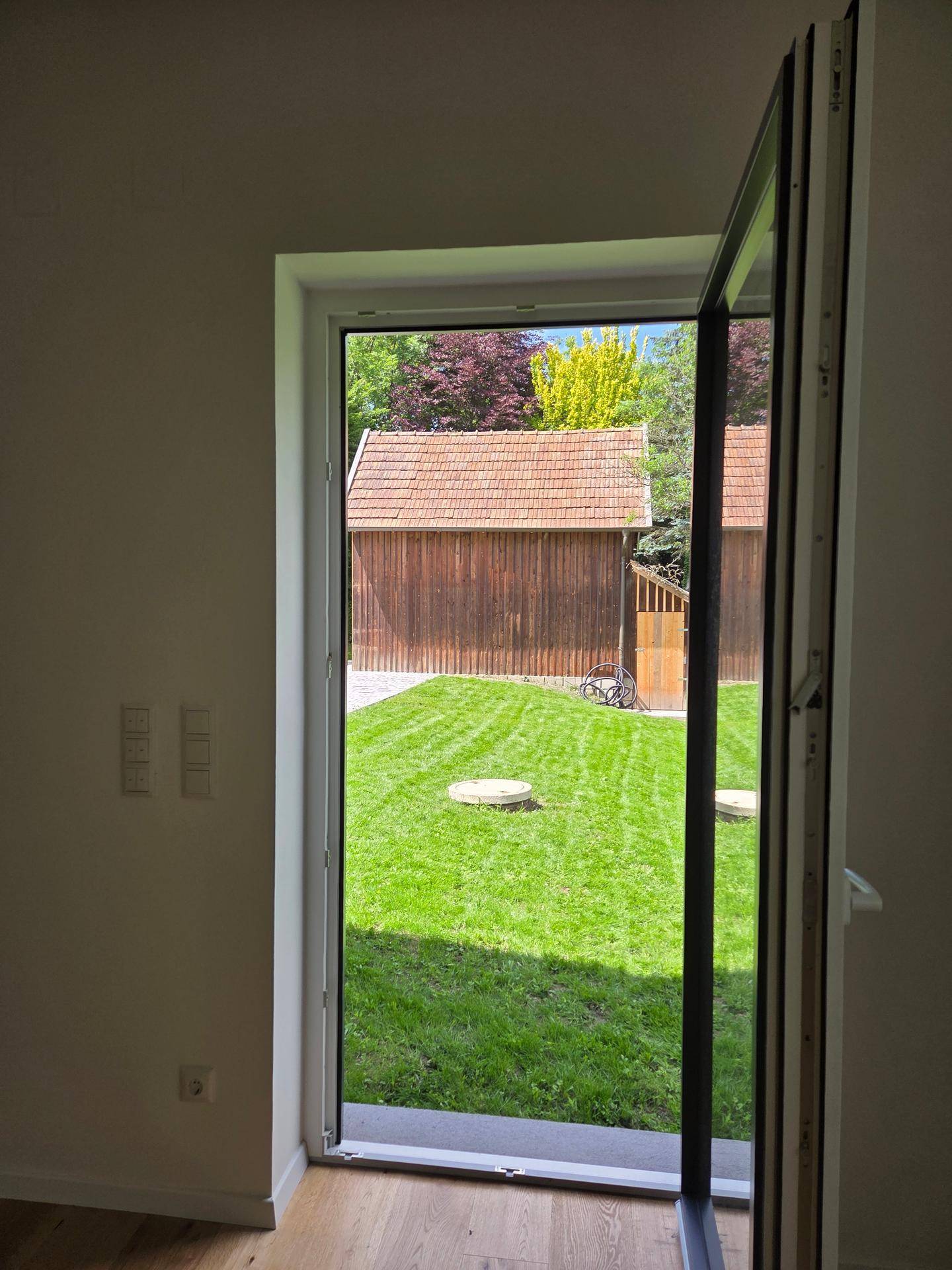 Blick aus einem Zimmer auf den grünen Garten mit einer Holzhütte im Hintergrund.