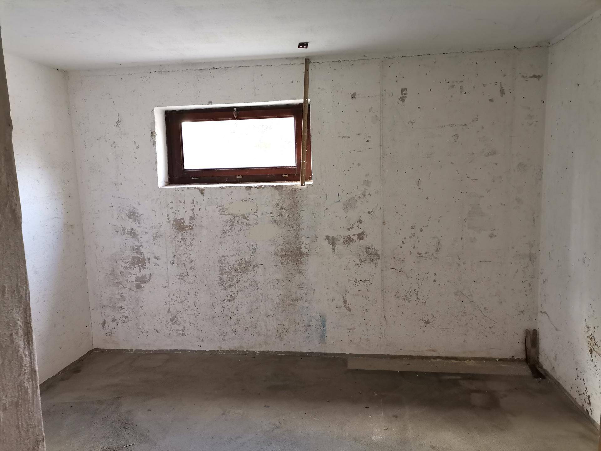 Kellerraum mit kleinem Fenster, Betonboden und unfertigen Wänden.