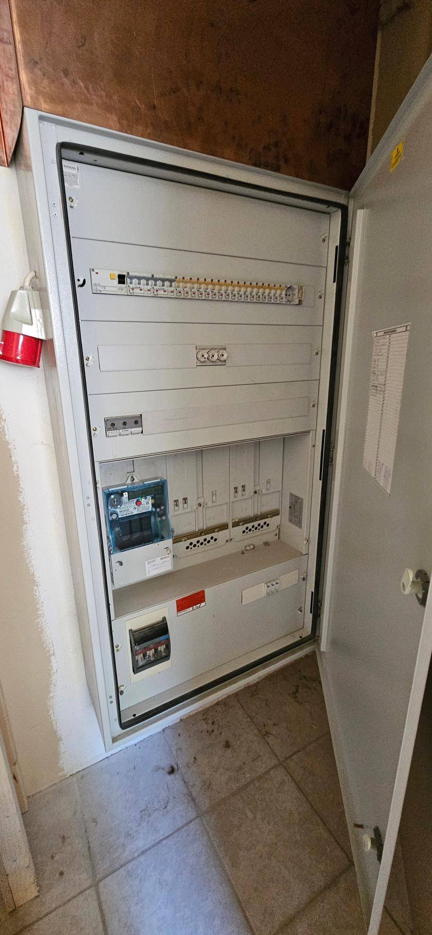 Offener Sicherungskasten in einem Technikraum mit Fliesenboden und einfacher Ausstattung.