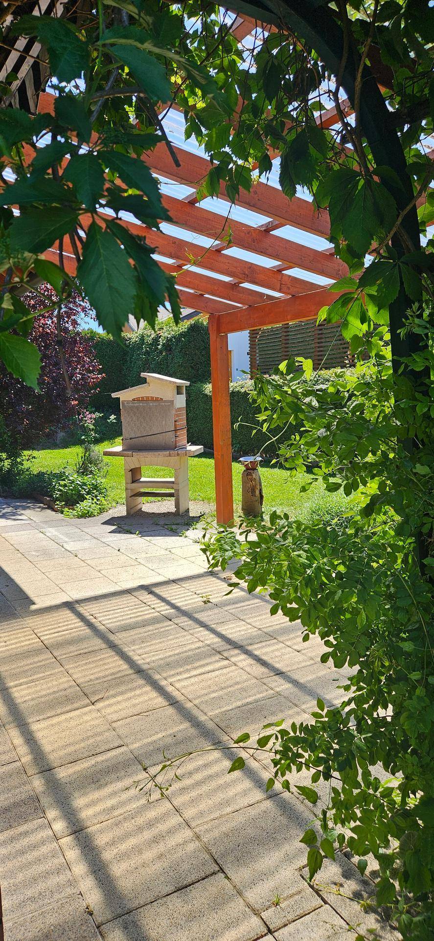 Gepflasterte Terrasse unter einer Holzpergola, umgeben von grünen Pflanzen.