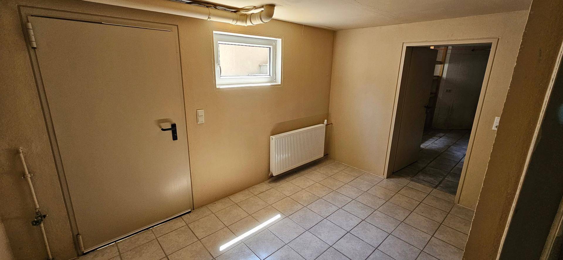 Einfacher Raum im Untergeschoss mit Fliesenboden, kleinem Fenster und Heizkörper.