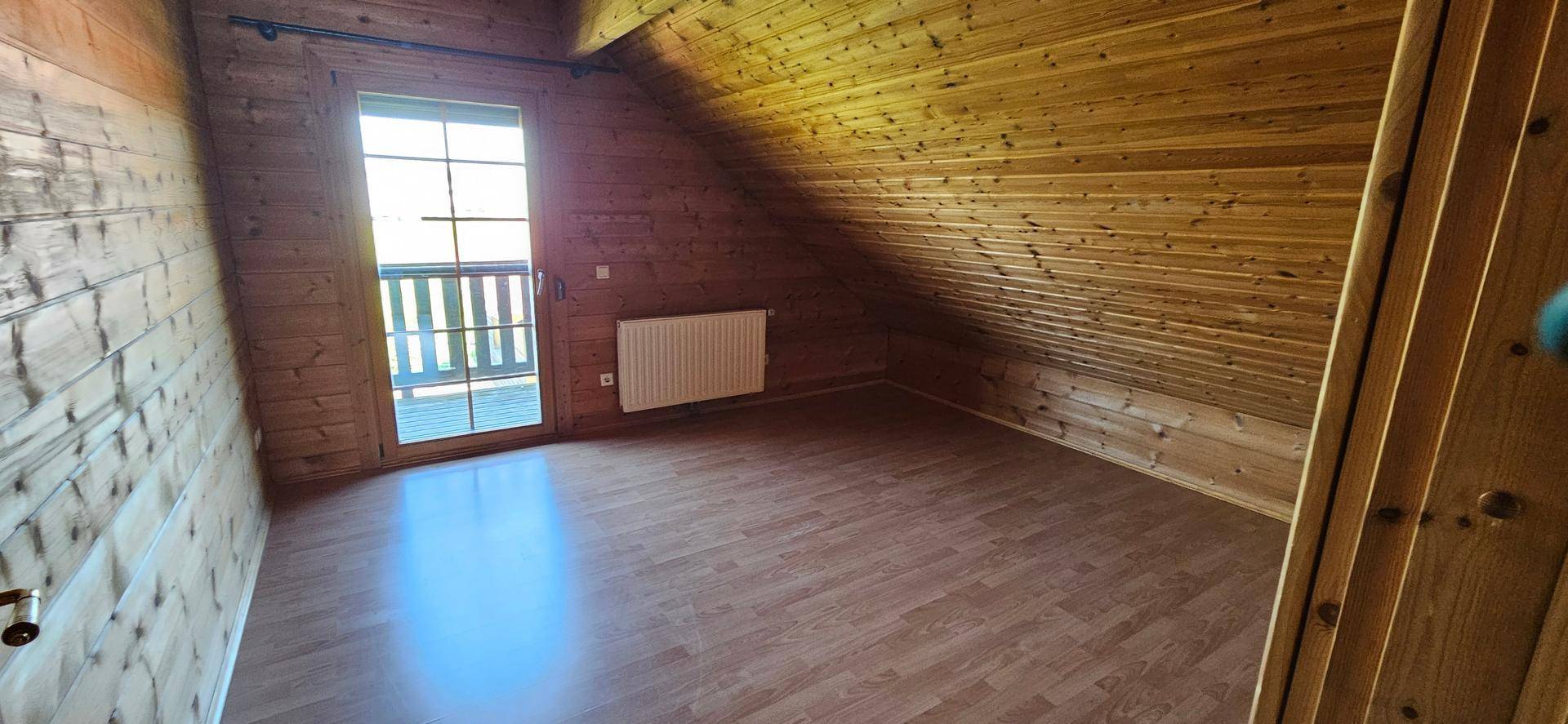 Dachgeschosszimmer mit Holzvertäfelung, Laminatboden und Zugang zu einem kleinen Balkon.