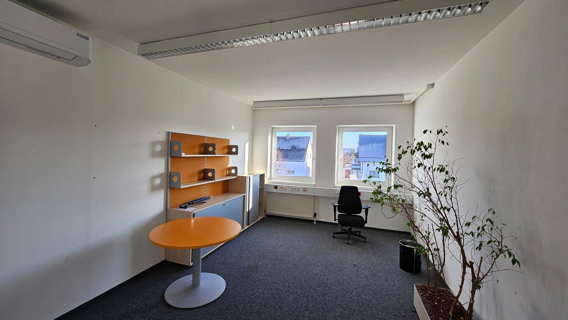 Büro mit Teppichboden, Schreibtisch und Pflanzen, sowie Fenstern und Klimaanlage.