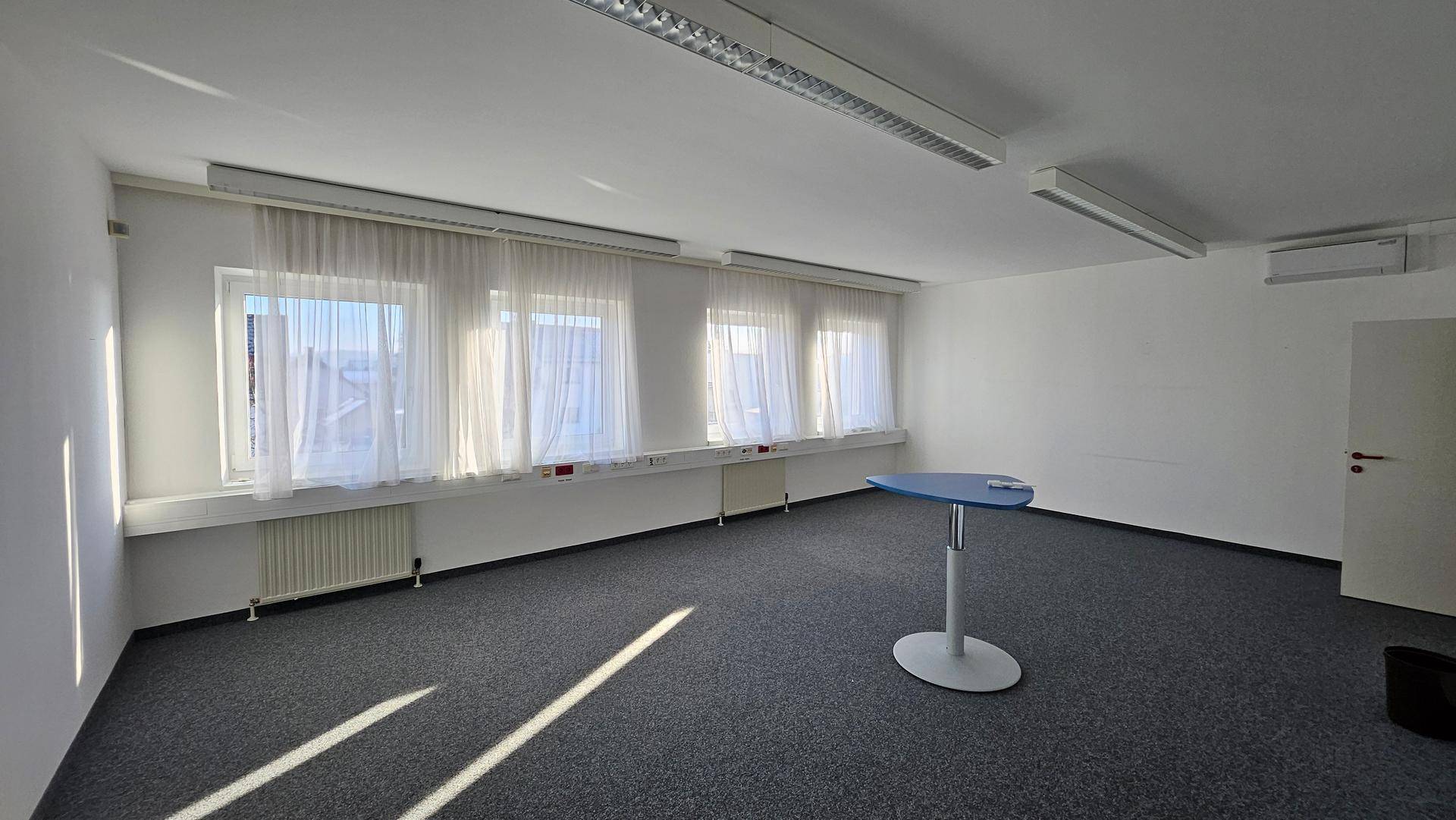 Helles Büro mit mehreren Fenstern, Teppichboden und einem kleinen Stehtisch, viel Tageslicht.