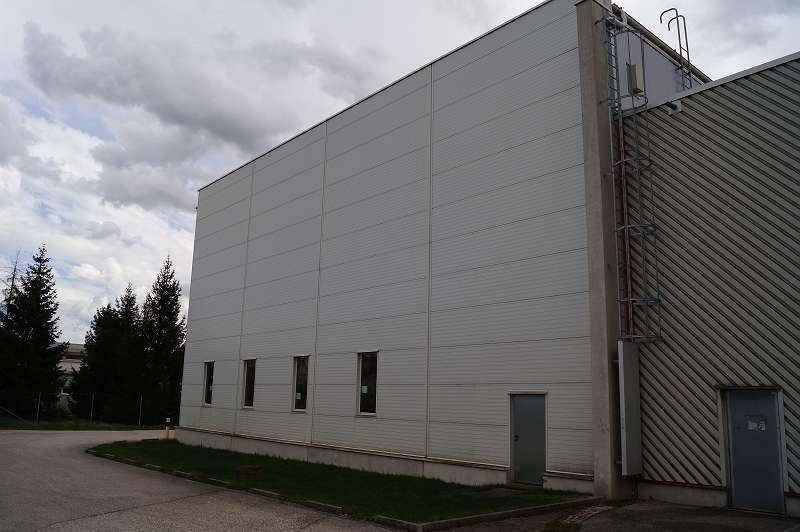 Seitenansicht einer großen Lagerhalle mit Metallfassade und kleinen Fenstern unter bewölktem Himmel.
