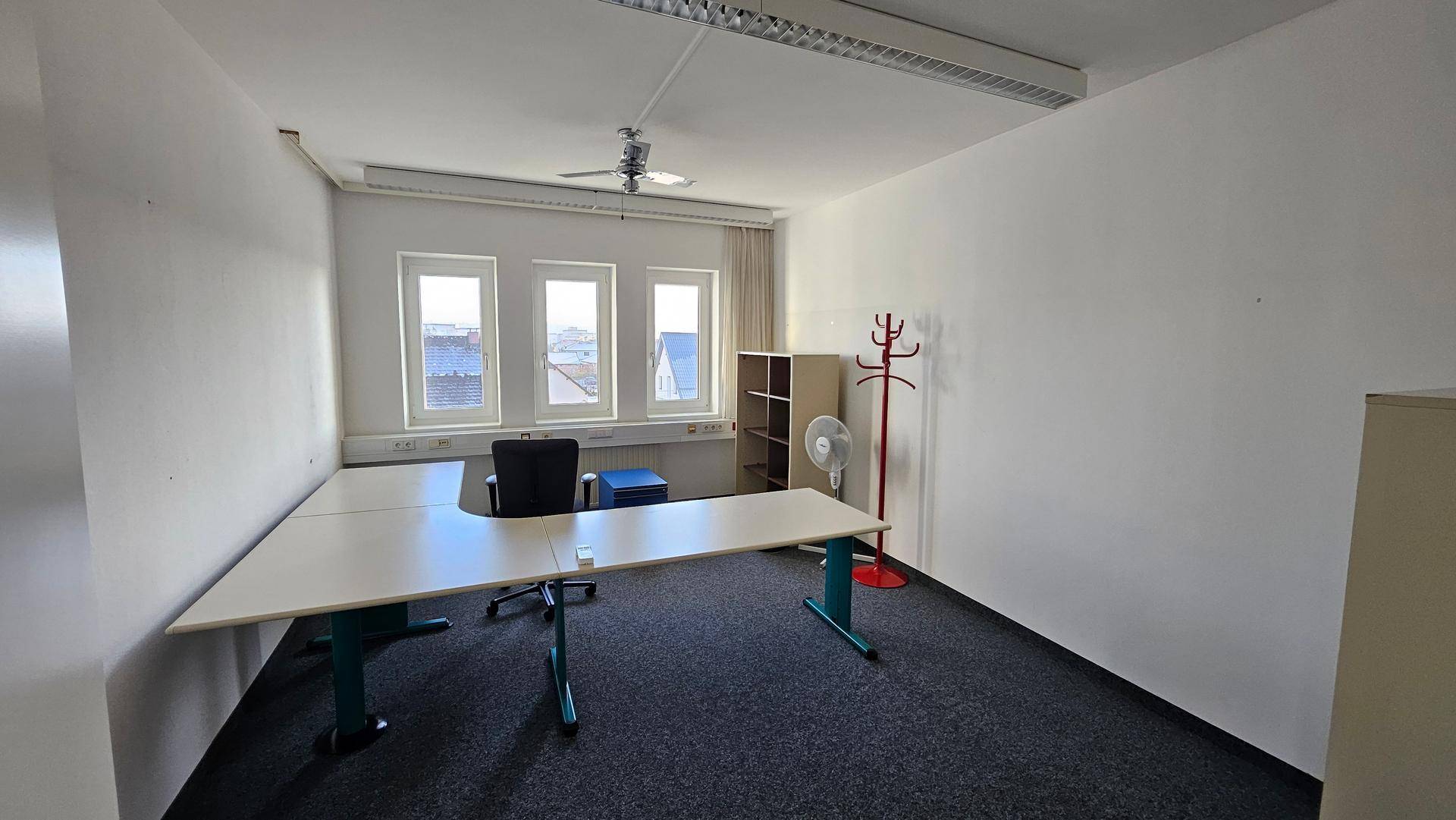 Büro mit Teppichboden, Schreibtisch und Stuhl, sowie einem Deckenventilator und Fenstern.