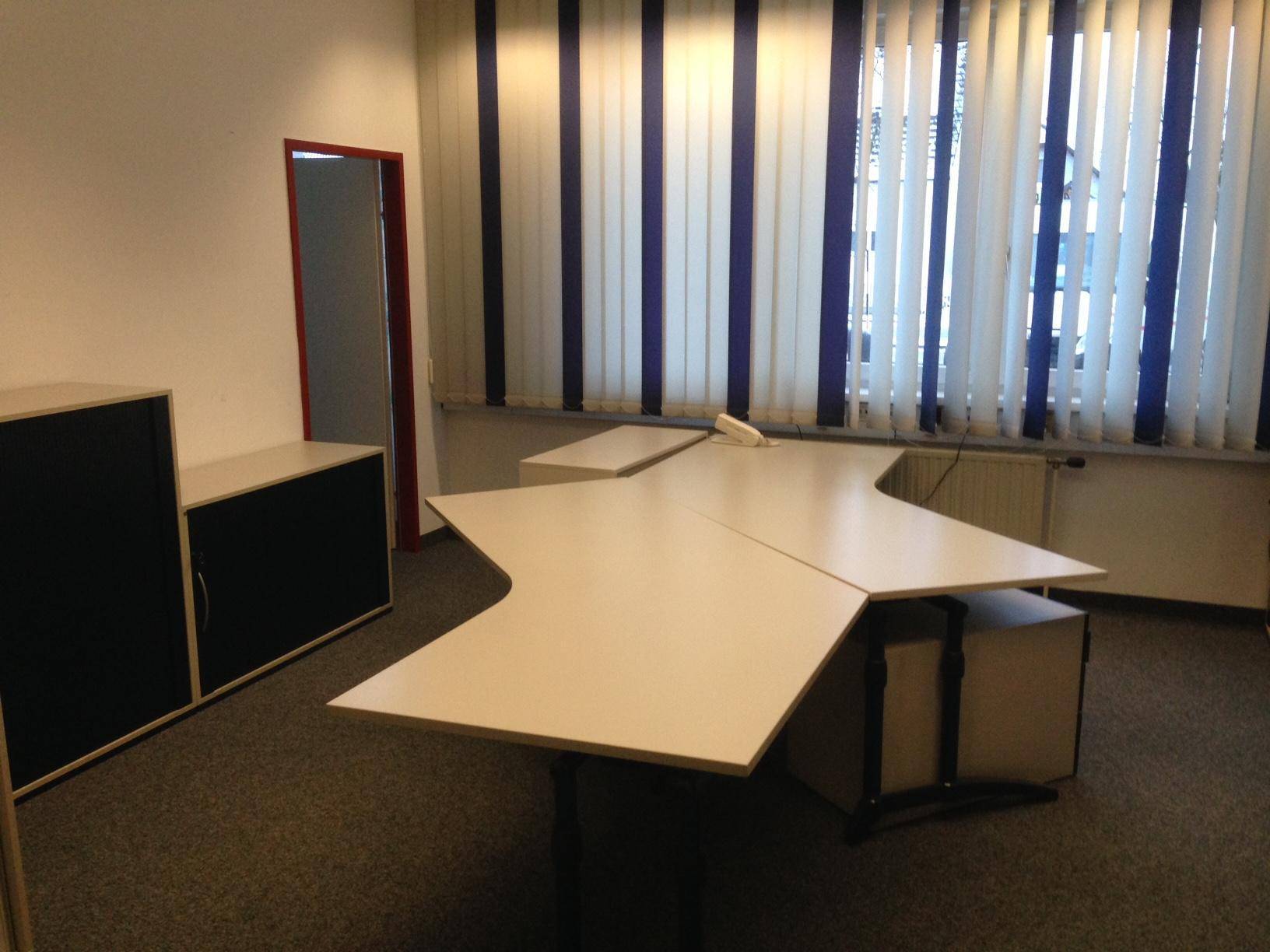 Büro mit Teppichboden, Schreibtisch und Schränken, sowie vertikalen Jalousien an den Fenstern.