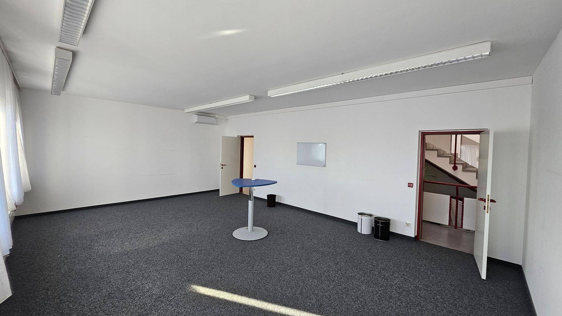 Helles Büro mit Teppichboden, weißen Wänden und einem kleinen Stehtisch, bereit zur Nutzung.