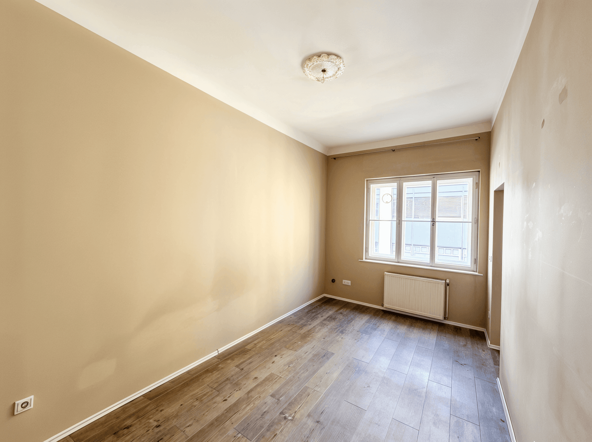 Helles, unmöbliertes Zimmer mit Holzboden, großem Fenster und Heizkörper.