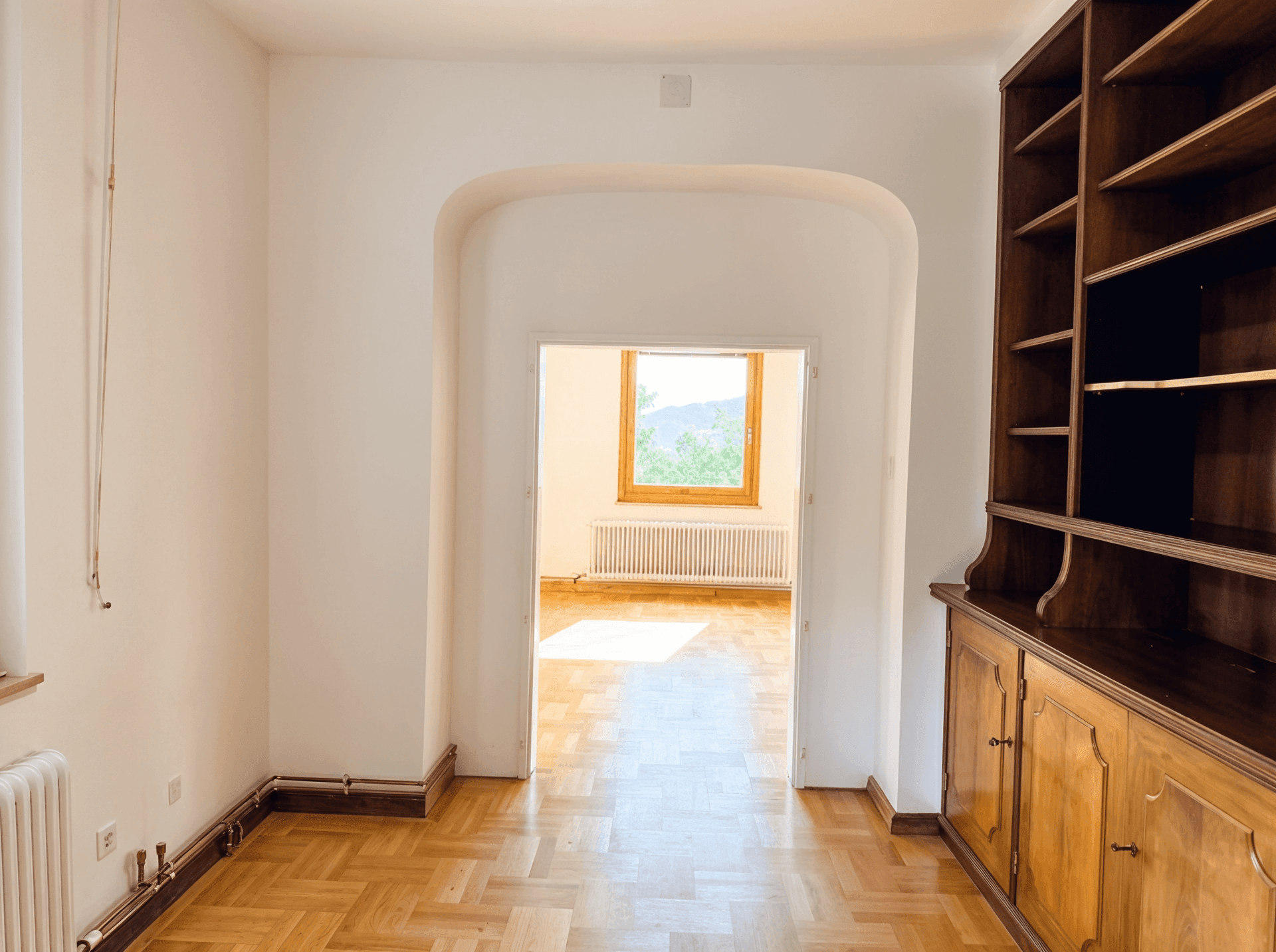 Heller Raum mit Parkettboden und einem großen Holzbücherregal, verbunden durch einen Bogen.