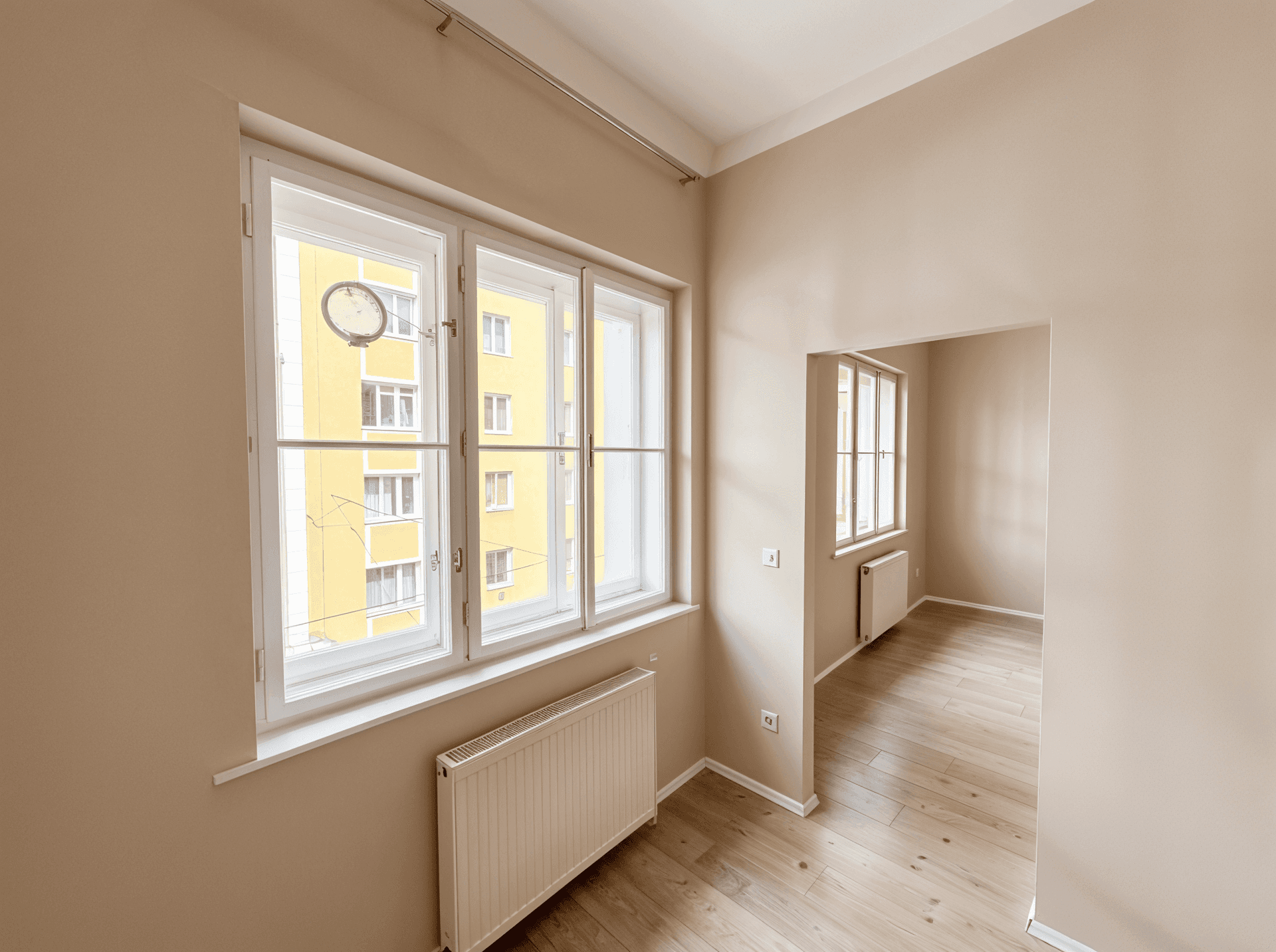 Großes Fenster in einem hellen Zimmer mit Holzboden und Blick auf ein gegenüberliegendes Gebäude.