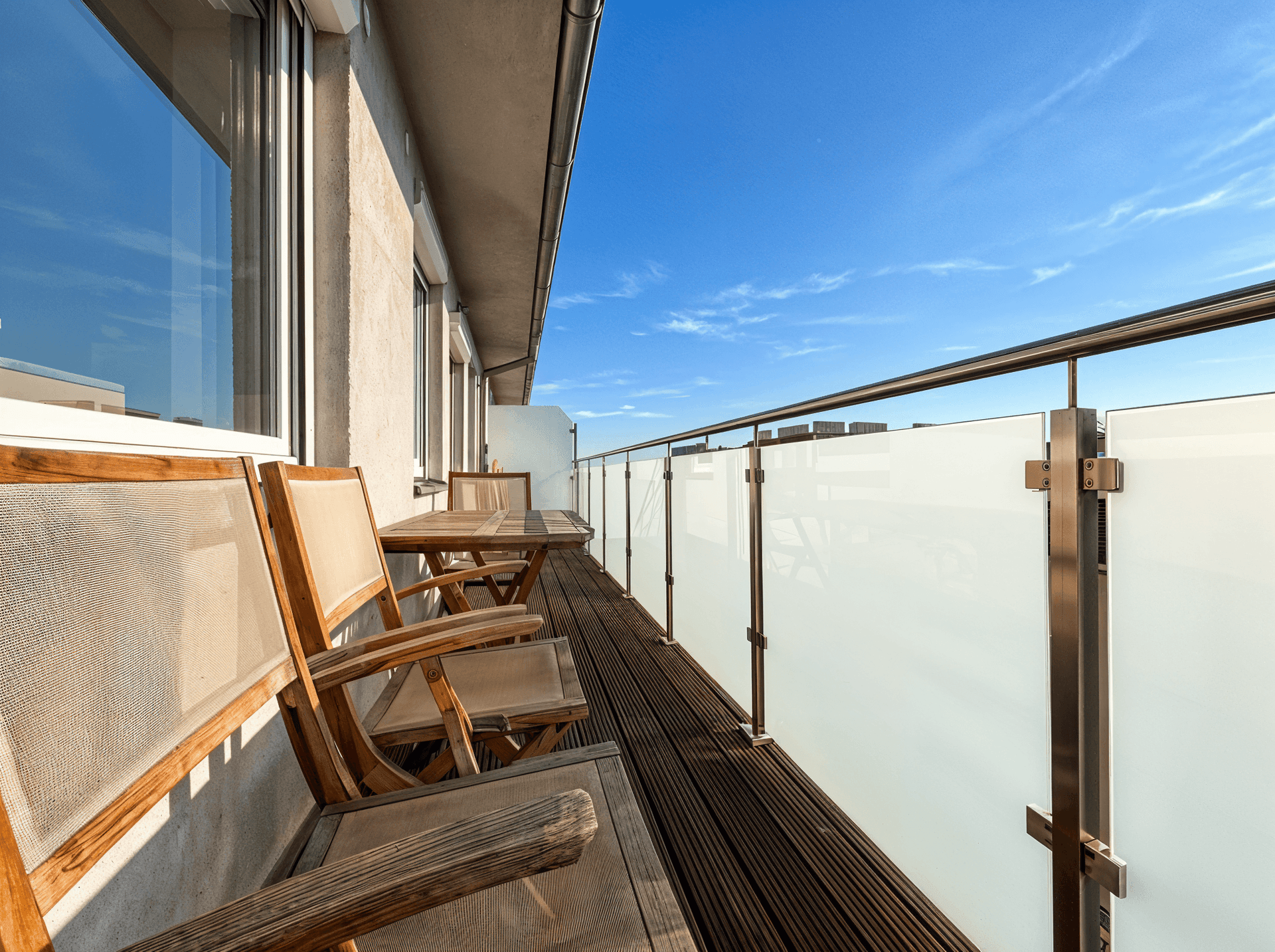 Geräumiger Balkon mit Holzdeck, Glasgeländer und Gartenmöbeln unter blauem Himmel.