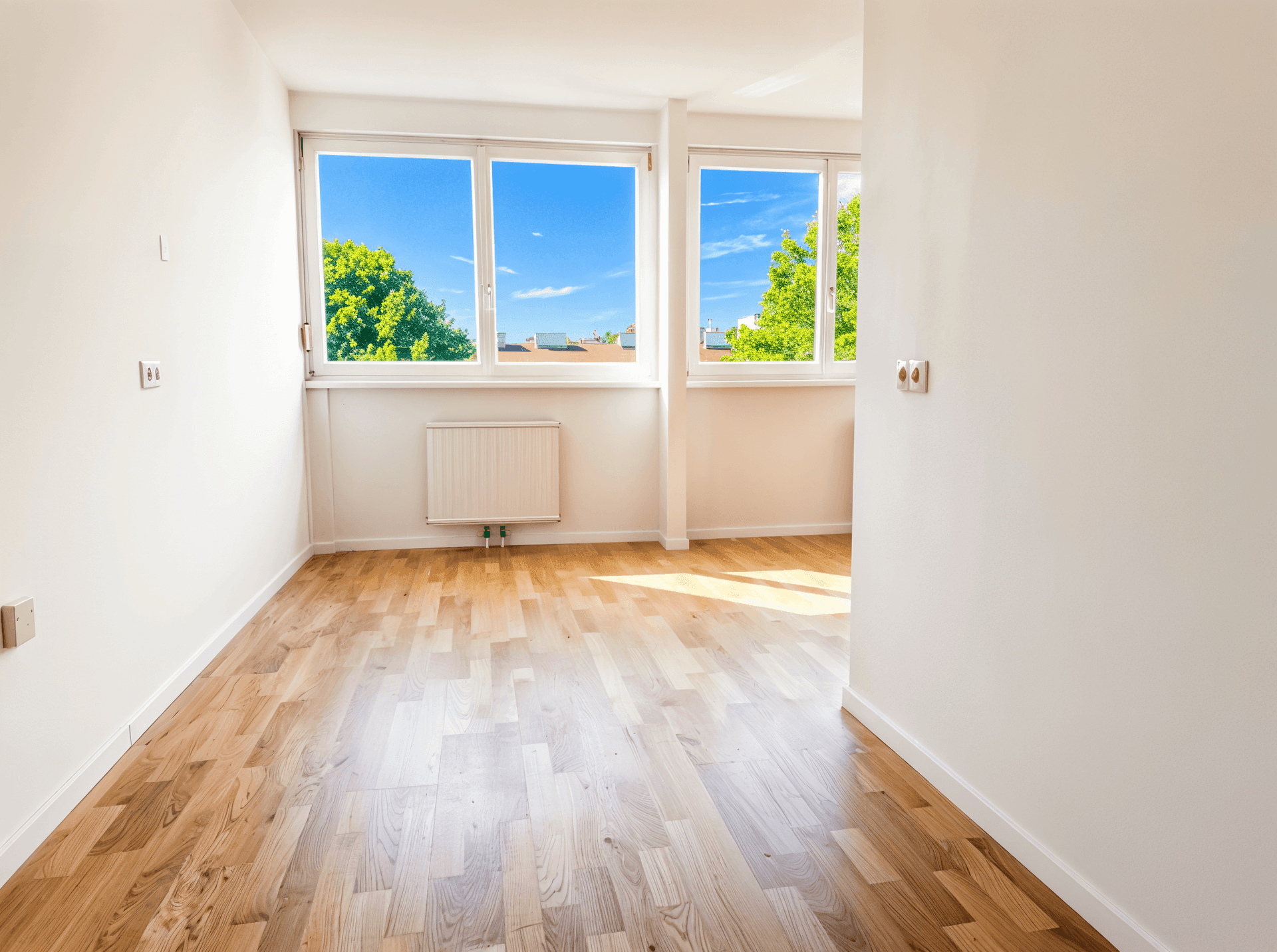 Heller, leerer Raum mit Holzboden und großen Fenstern mit Blick ins Grüne.