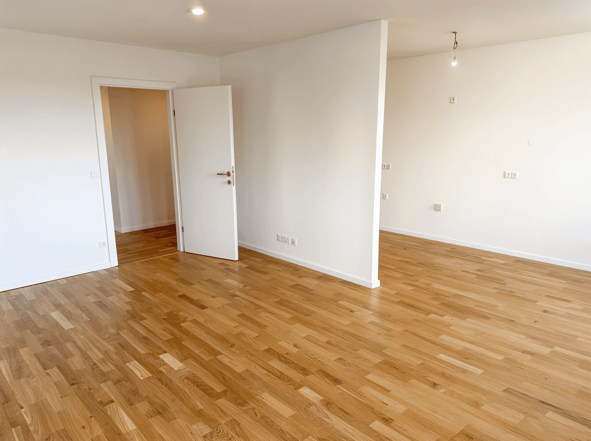 Leerer Raum mit Holzboden und weiß gestrichenen Wänden, mit Zugang zu einem weiteren Zimmer.
