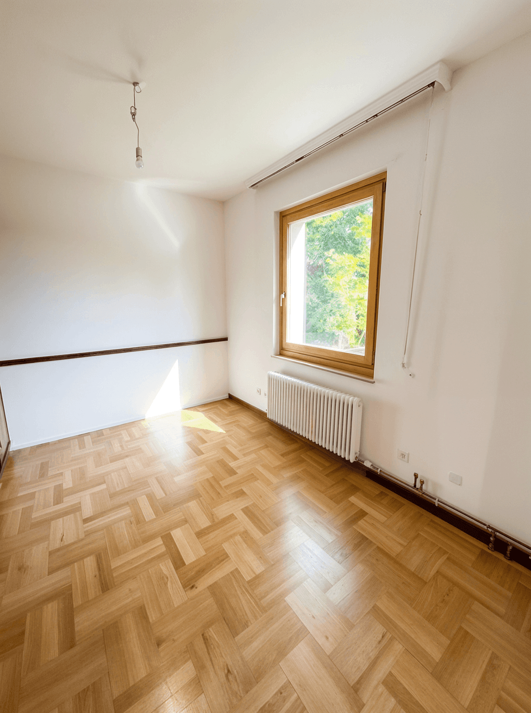 Kleiner, heller Raum mit Fischgrätparkett und einem Fenster mit Blick ins Grüne.