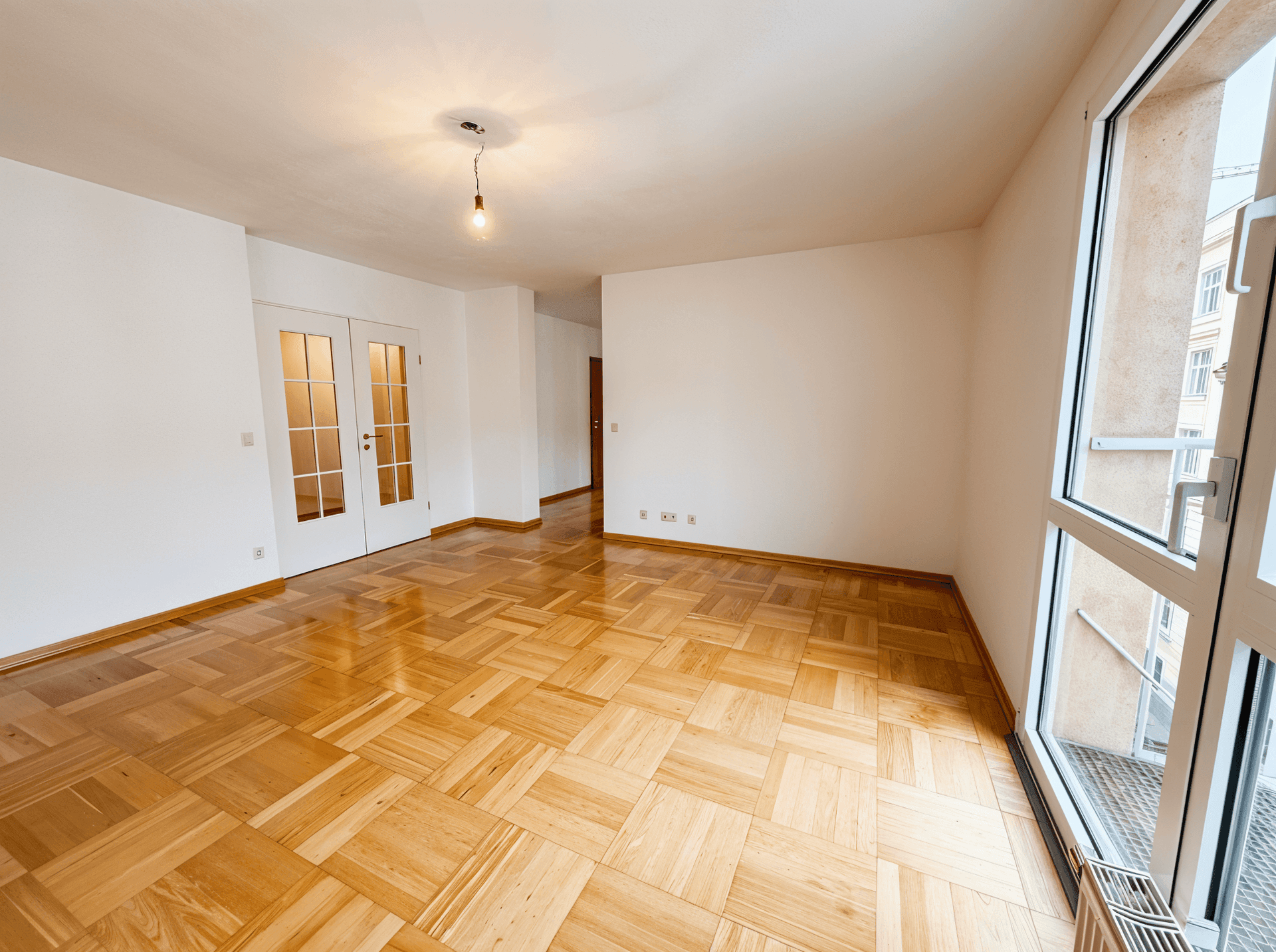 Geräumiges, leeres Zimmer mit Parkettboden und großen Fenstern, die viel Licht hereinlassen.