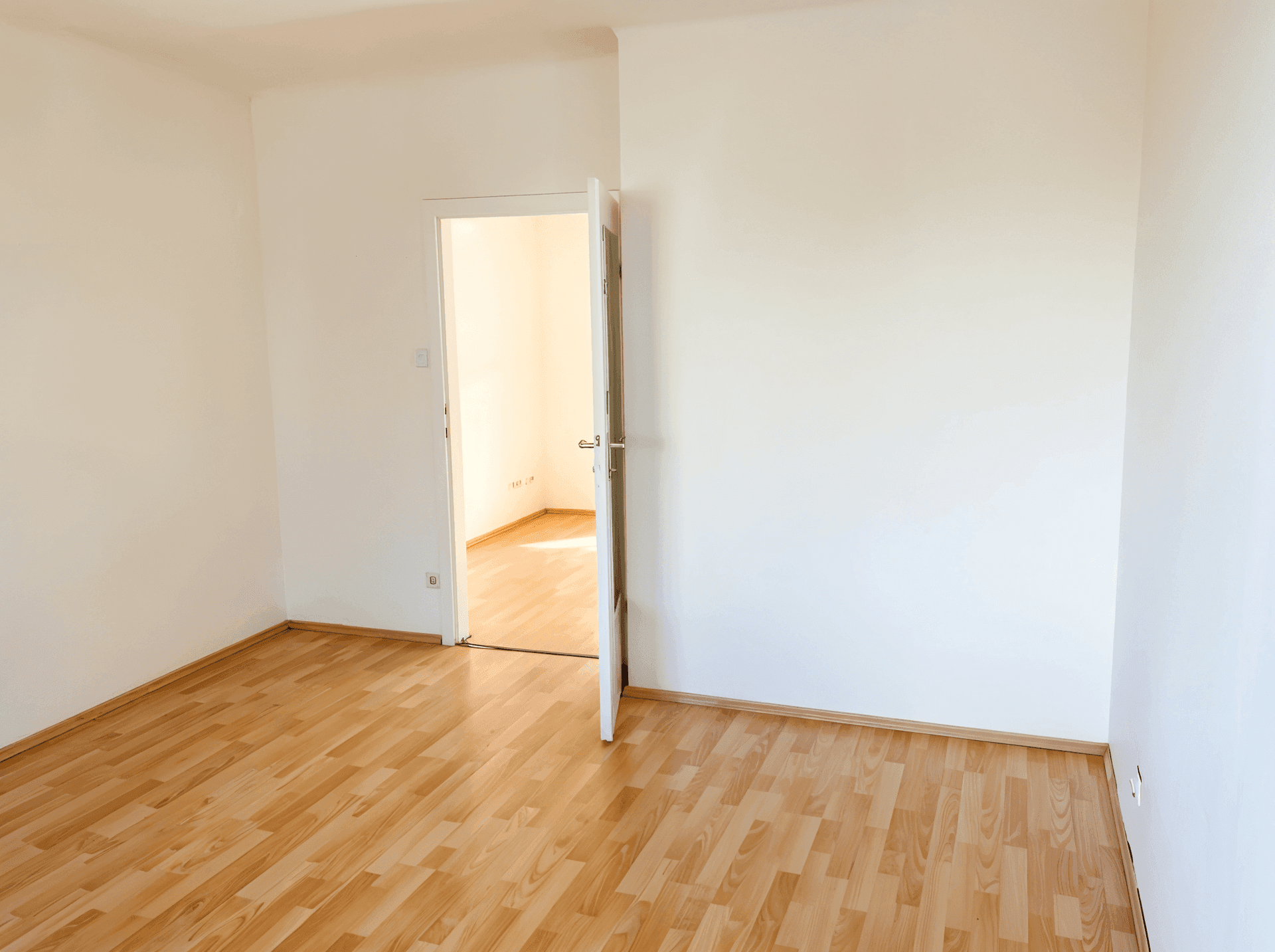 Leerer Raum mit hellem Parkettboden und weiß gestrichenen Wänden, der Zugang zu einem weiteren Zimmer bietet.