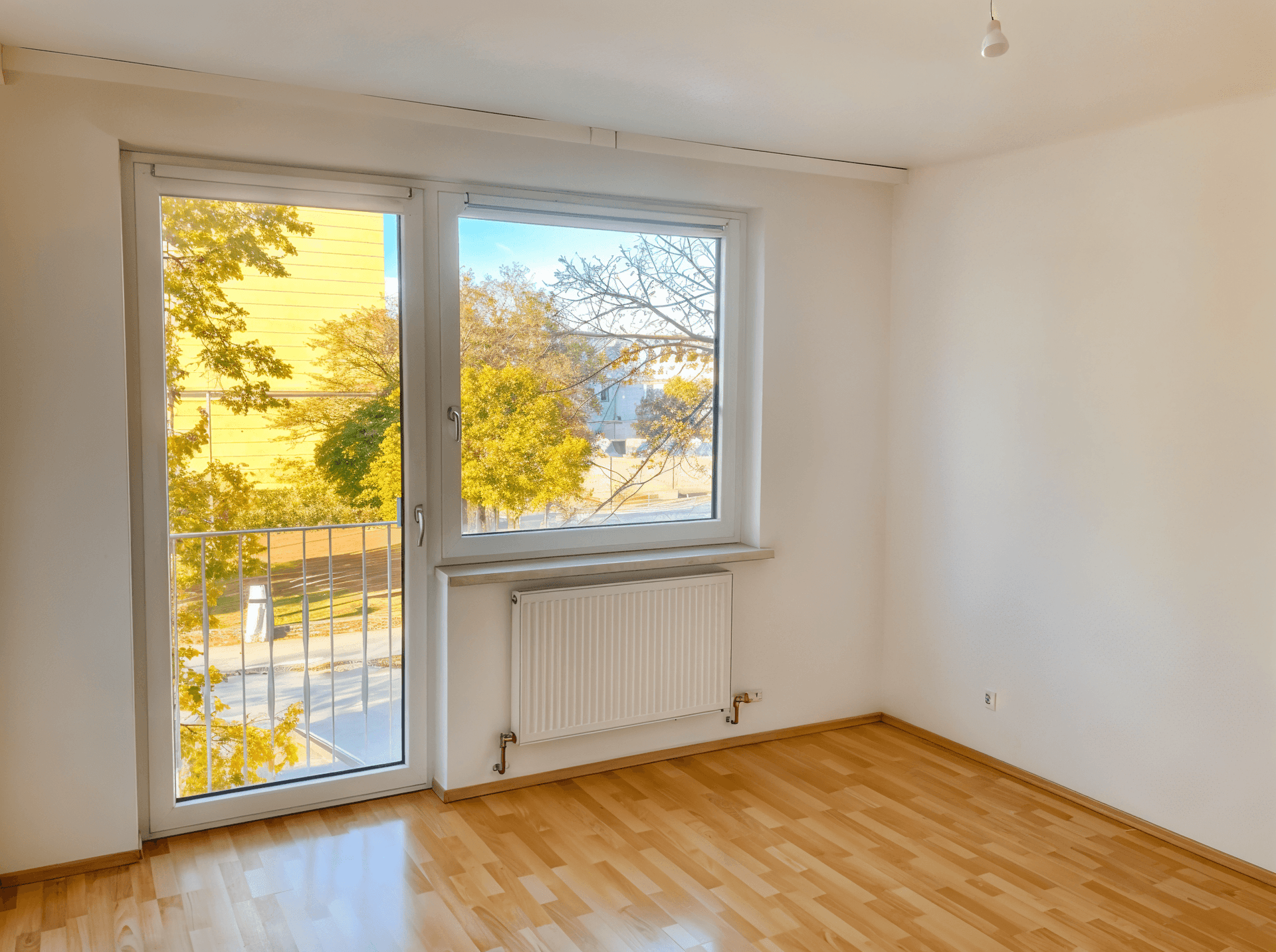 Lichtdurchfluteter Raum mit Holzboden und Blick ins Grüne, ausgestattet mit einem Heizkörper unter dem Fenster.